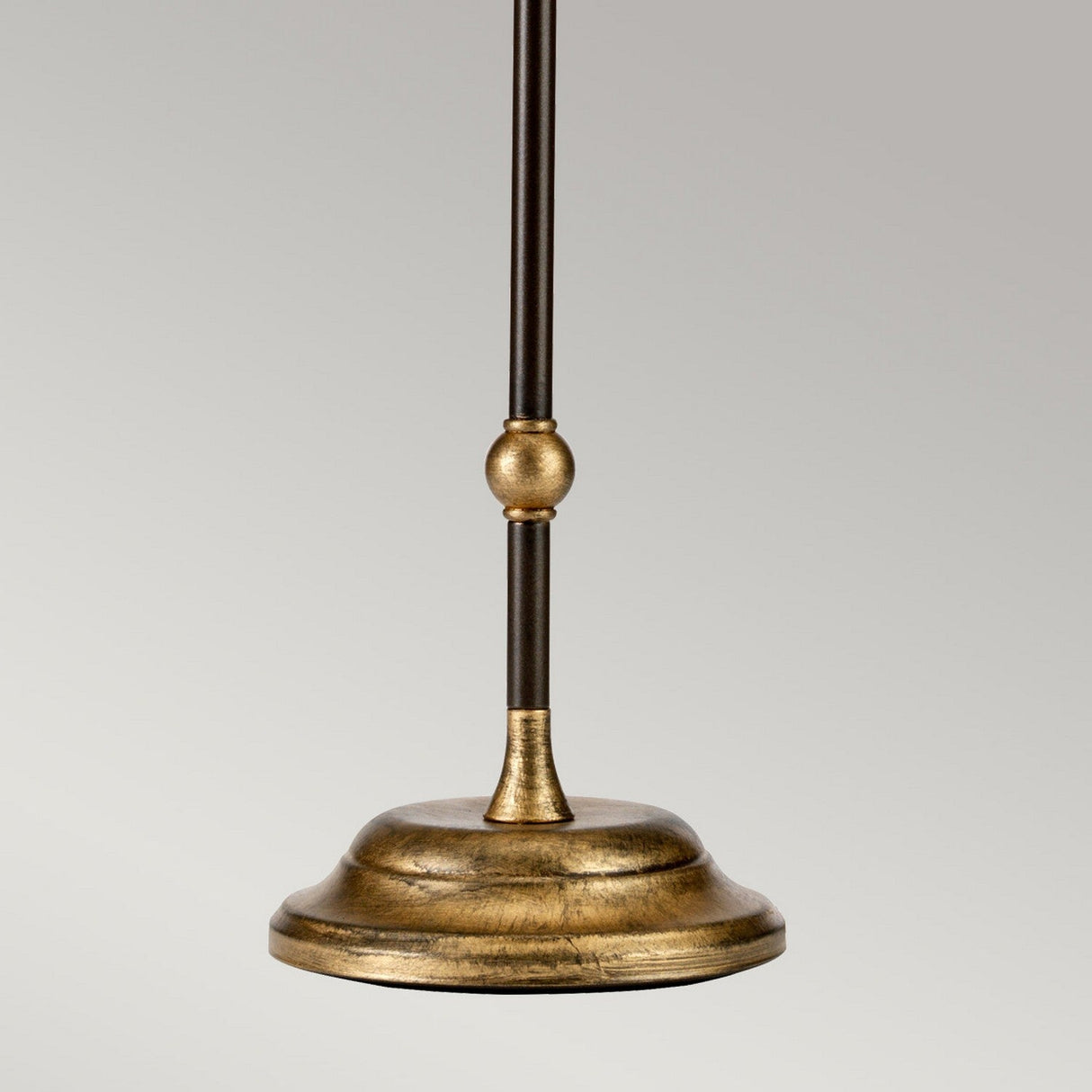 A detailed view of the Amarilli 1 Light Table Lamp showcases its brass pendant light fixture with a bronze finish and gold patina. This handmade lamp features a slim rod adorned with a decorative orb at its center, which extends to a round base set against a neutral gray background.