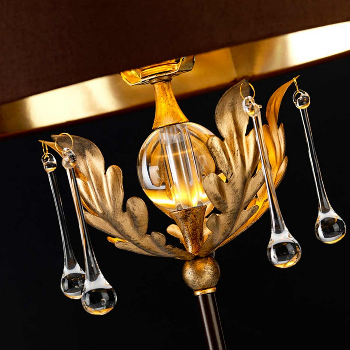 A close-up of the Amarilli 1 Light Table Lamp showcases a transparent central sphere embellished with bronze accents featuring a gold patina and adorned with hanging glass droplets. The warm lighting emits a golden glow, accentuating the intricate details against the dark background.