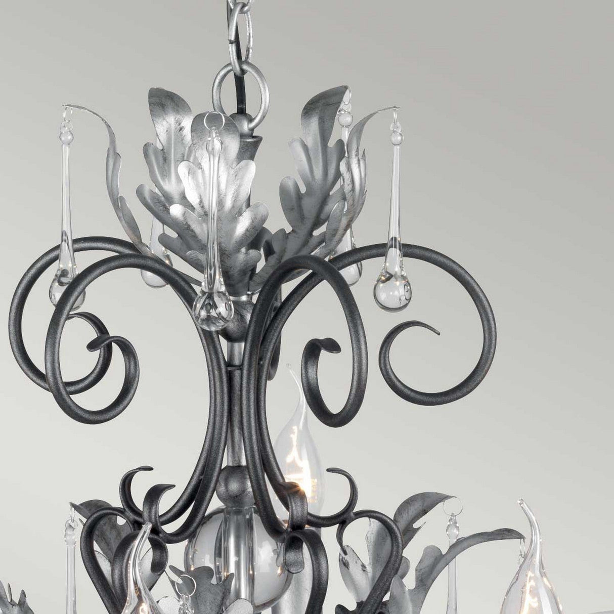 A close-up of the Amarilli 10 Light Chandelier - Black With Silver Patina, highlighting its ornate swirling metalwork and decorative teardrop-shaped glass pendants. The piece's elegant finish is set against a plain light gray background, accentuating its sophistication.