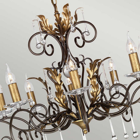 The Amarilli 10 Light Chandelier - Bronze With Gold Patina is an elegant piece featuring ornate metallic scrollwork and multiple candle-like bulbs adorned with decorative crystal drops, all set against a light gray background, making it the perfect luxurious lighting fixture.