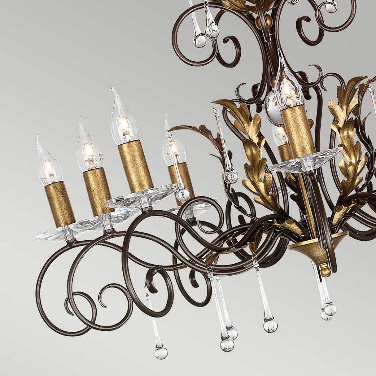 The Amarilli 10 Light Chandelier in Bronze with Gold Patina is a luxurious lighting fixture featuring intricate metalwork and candle-shaped bulbs. Crystal drops dangle elegantly from its arms, adding an elegant touch that enhances any space against a light background.