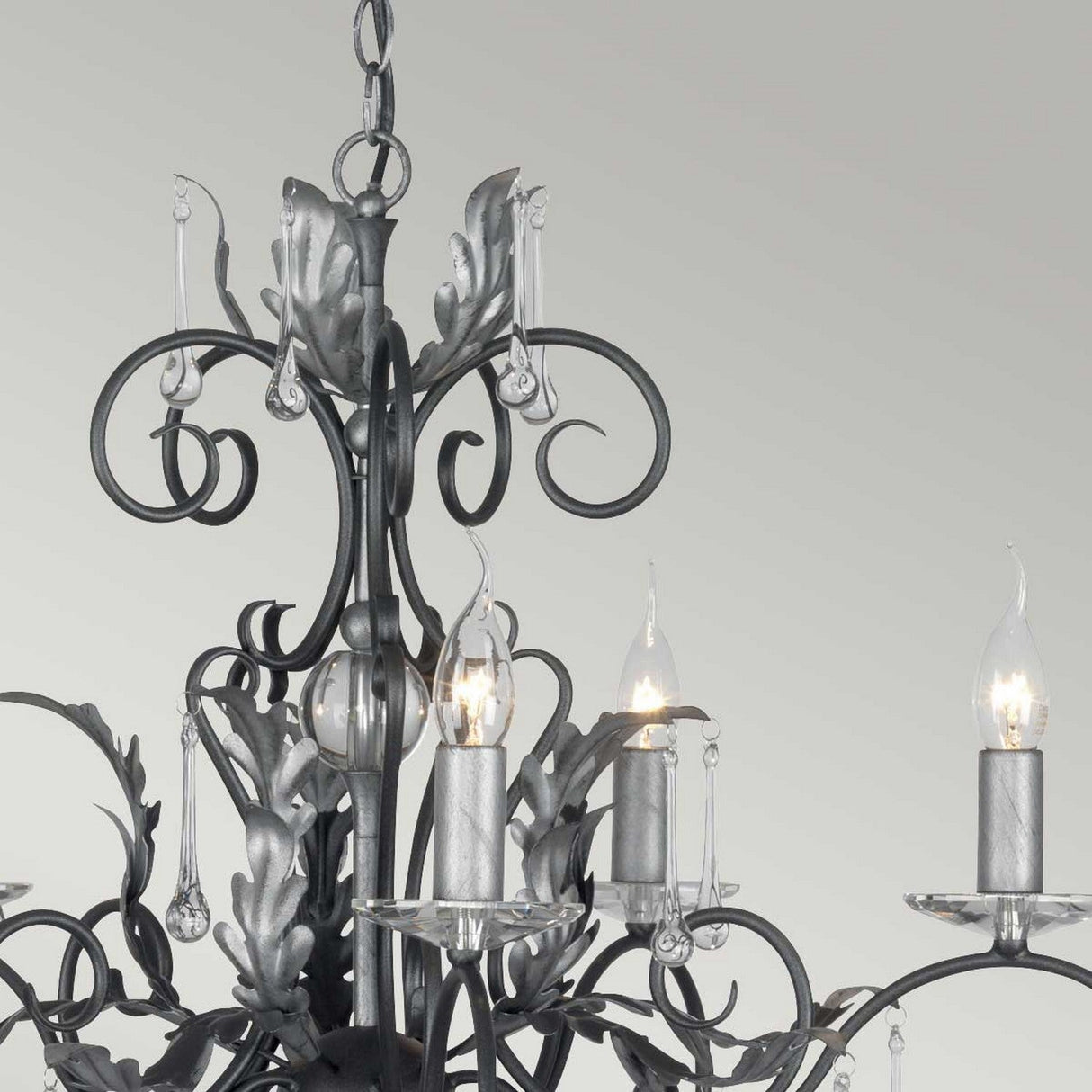 A close-up shot of the Amarilli 15 Light Chandelier reveals its ornate black metal with silver patina. The chandelier's decorative scrollwork and leaf accents perfectly complement the candle-like bulb holders, each featuring a flame-shaped light. A neutral gray backdrop enhances the intricate design details of this stunning piece.