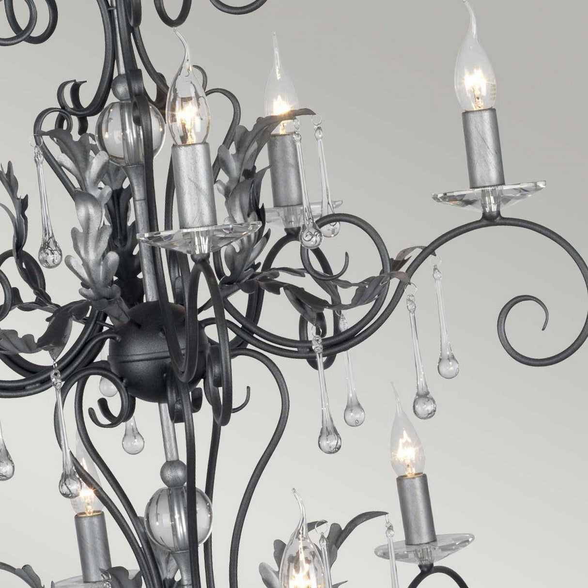 The Amarilli 15 Light Chandelier in black with silver patina showcases a detailed close-up of its elegant, classic design. This chandelier features numerous candle-shaped light bulbs, intricate metal scrollwork, and dangling crystal-like beads.