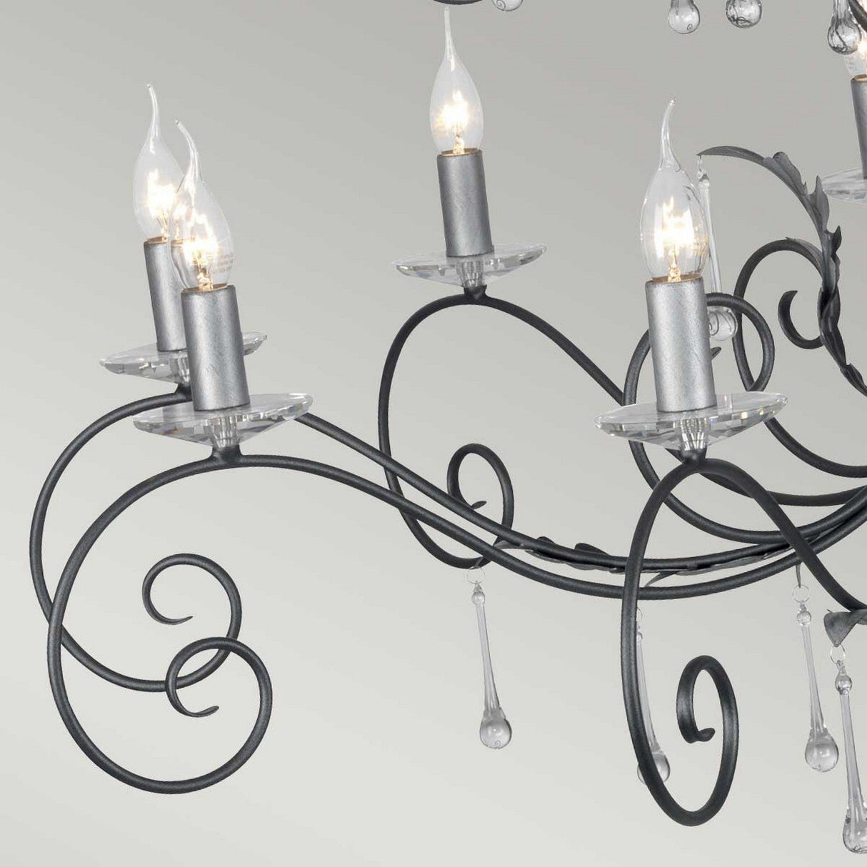 The Amarilli 15 Light Chandelier - Black With Silver Patina features intricate black metal scrollwork with a refined silver patina, beautifully outlining its exposed candle-style bulbs. The sophistication of the chandelier is further enriched by delicate glass droplets hanging from its structure.