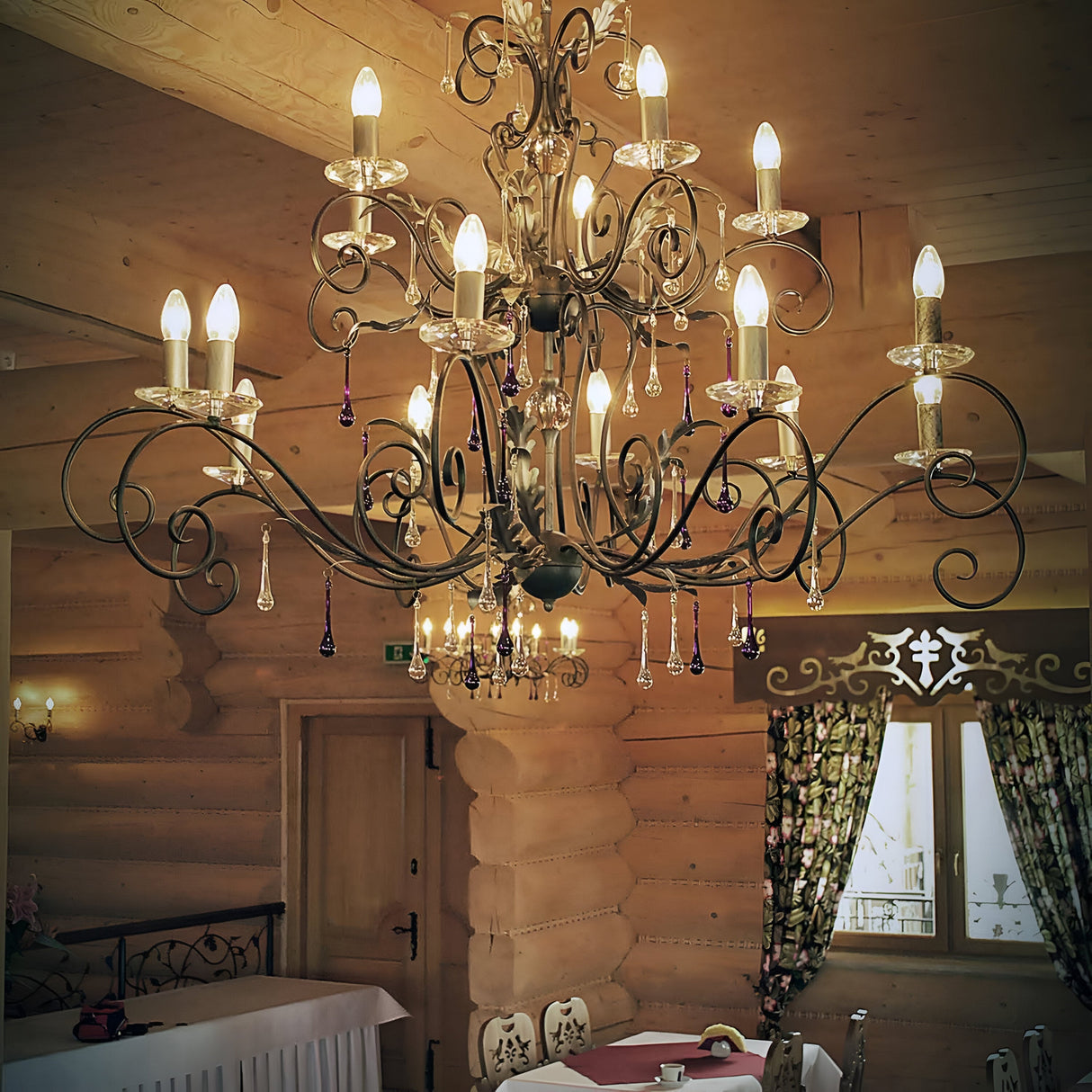 The Amarilli 15 Light Chandelier - Bronze With Gold Patina, a grand handmade fixture with multiple bulbs, illuminates the rustic wooden room. The space exudes coziness with its patterned curtains, wooden walls, and a neatly set table in the background.