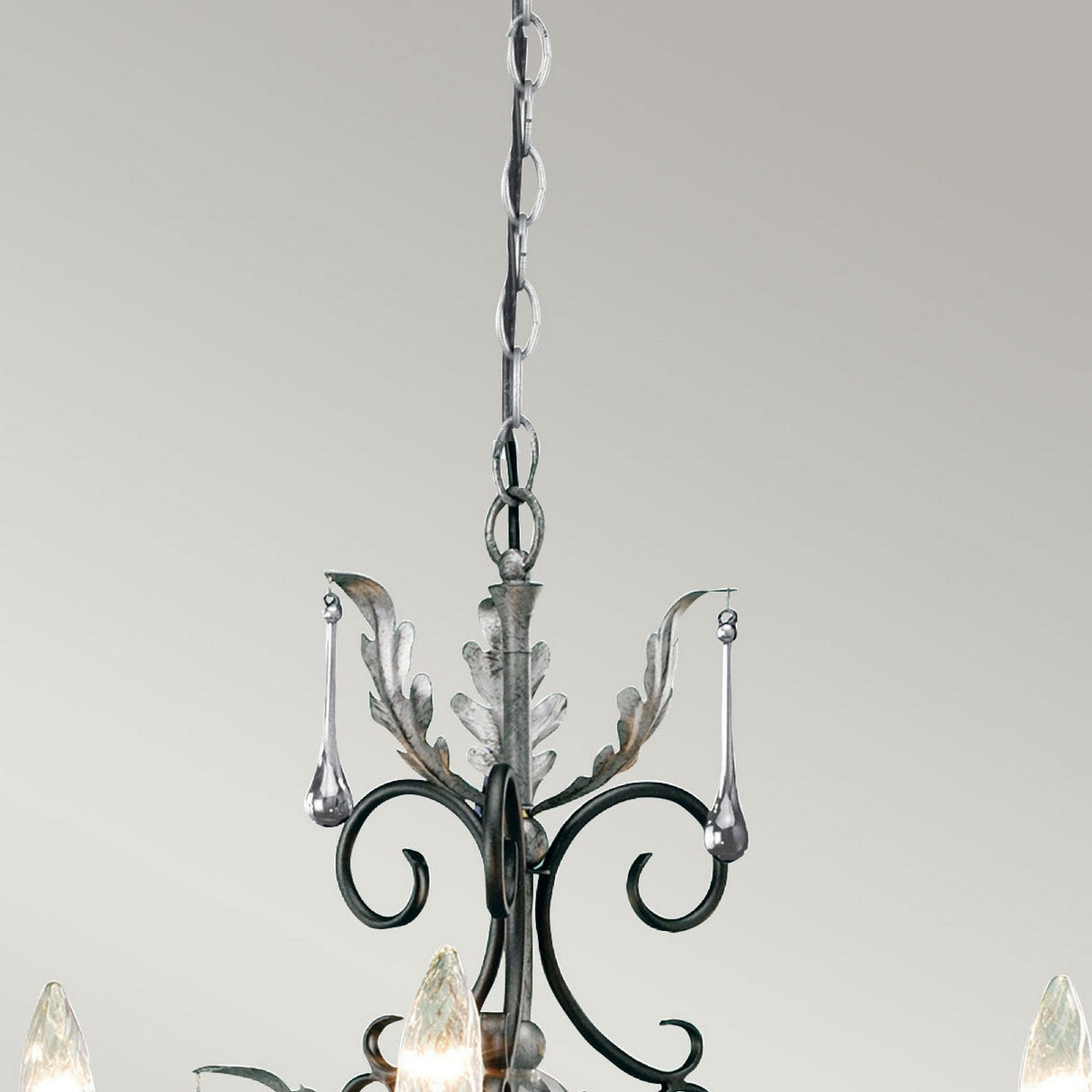A detailed view of the Amarilli 3 Light Chandelier - Black With Silver Patina showcases its intricate metal scrollwork and teardrop-shaped glass pendants. This elegant fixture, handcrafted by Elstead, is beautifully suspended by a chain against a light gray backdrop.