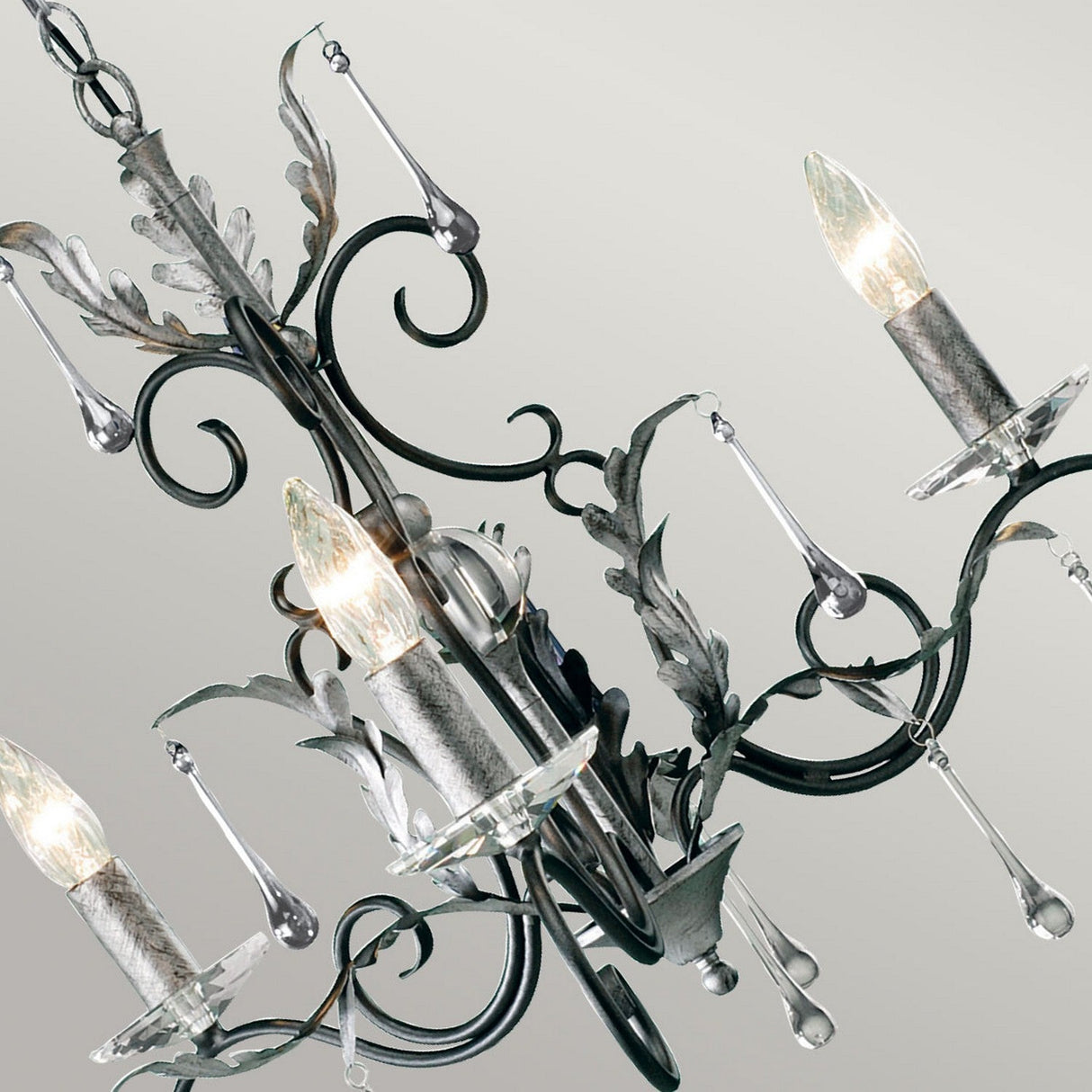 Here's a look at the Amarilli 3 Light Chandelier, expertly handcrafted by Elstead. The piece features detailed metalwork with graceful curves and leaf-inspired motifs, enhanced by a black finish with silver patina. It complements its three candle-like bulbs and dangling crystal ornaments beautifully against a light gray backdrop.