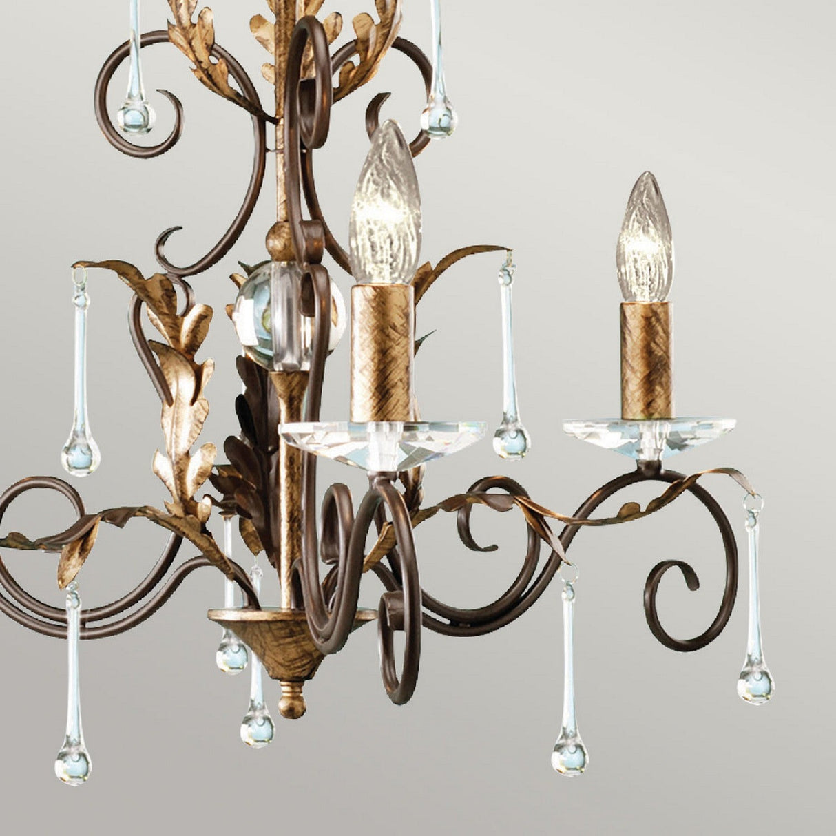 The Amarilli 3 Light Chandelier - Bronze With Gold Patina elegantly features two candle-like bulbs illuminated against a light gray backdrop. Its bronze metal arms, highlighted with a subtle gold patina, showcase intricate curves adorned with leaf motifs and teardrop-shaped crystals.