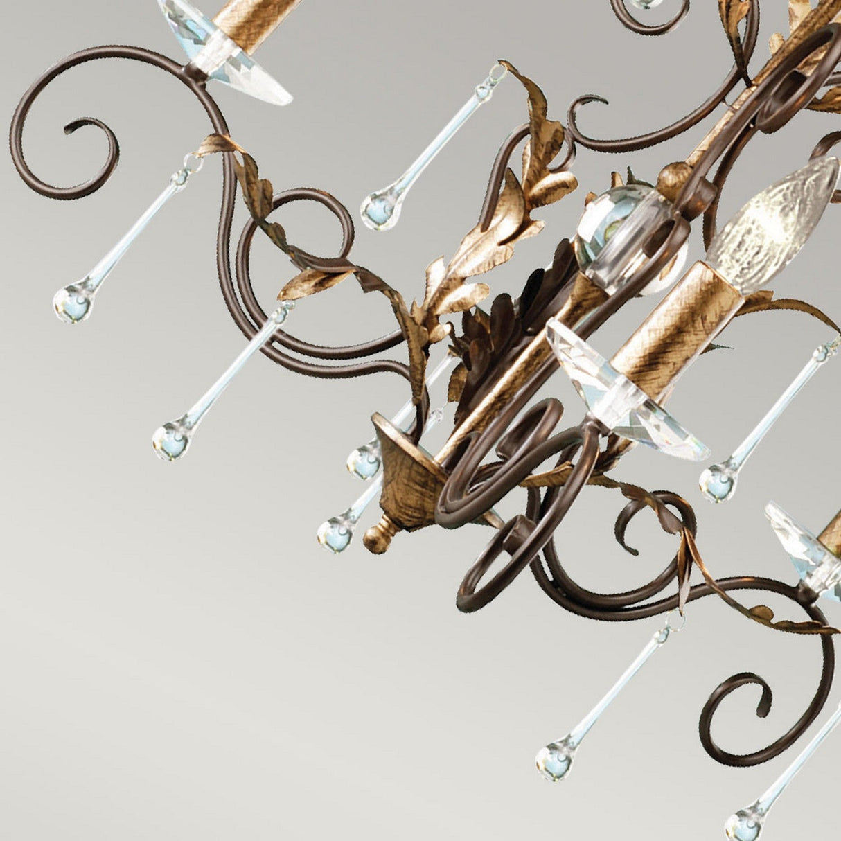 A close-up of the Amarilli 3 Light Chandelier highlights its vintage bronze with gold patina scrollwork and teardrop-shaped crystal pendants. This elegant lighting piece features candle-like bulb holders set against a plain light background, enhancing its timeless beauty.