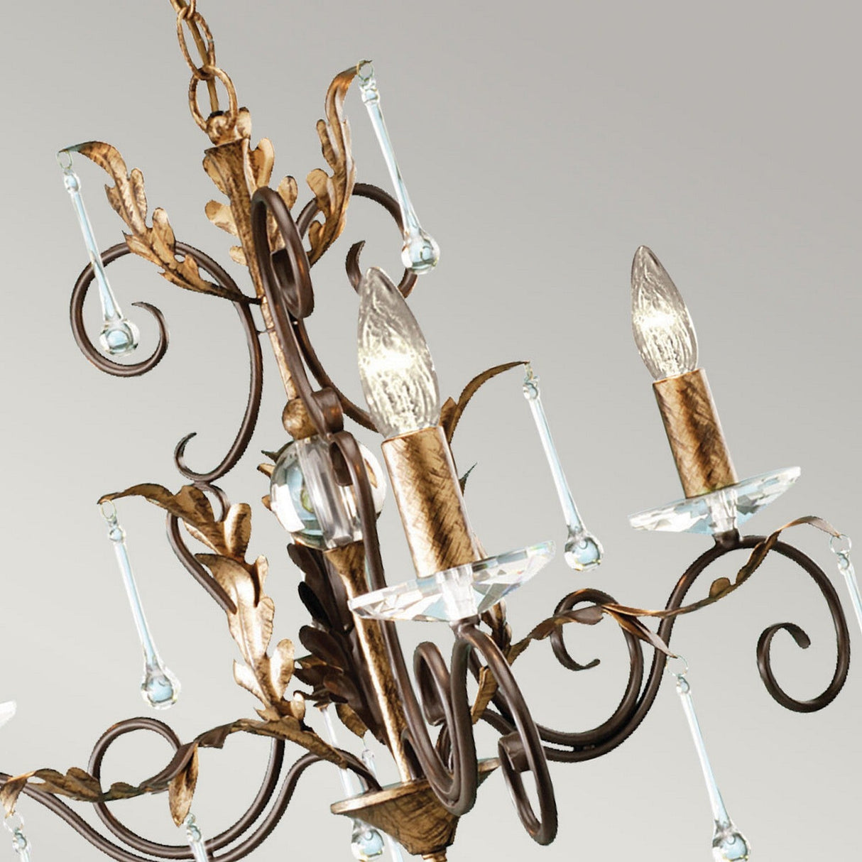 A detailed view of the Amarilli 3 Light Chandelier showcases its sophisticated bronze with gold patina design and clear crystal accents. The chandelier is adorned with candle-like bulbs and decorative droplets hanging from its branches, all set against a neutral background.