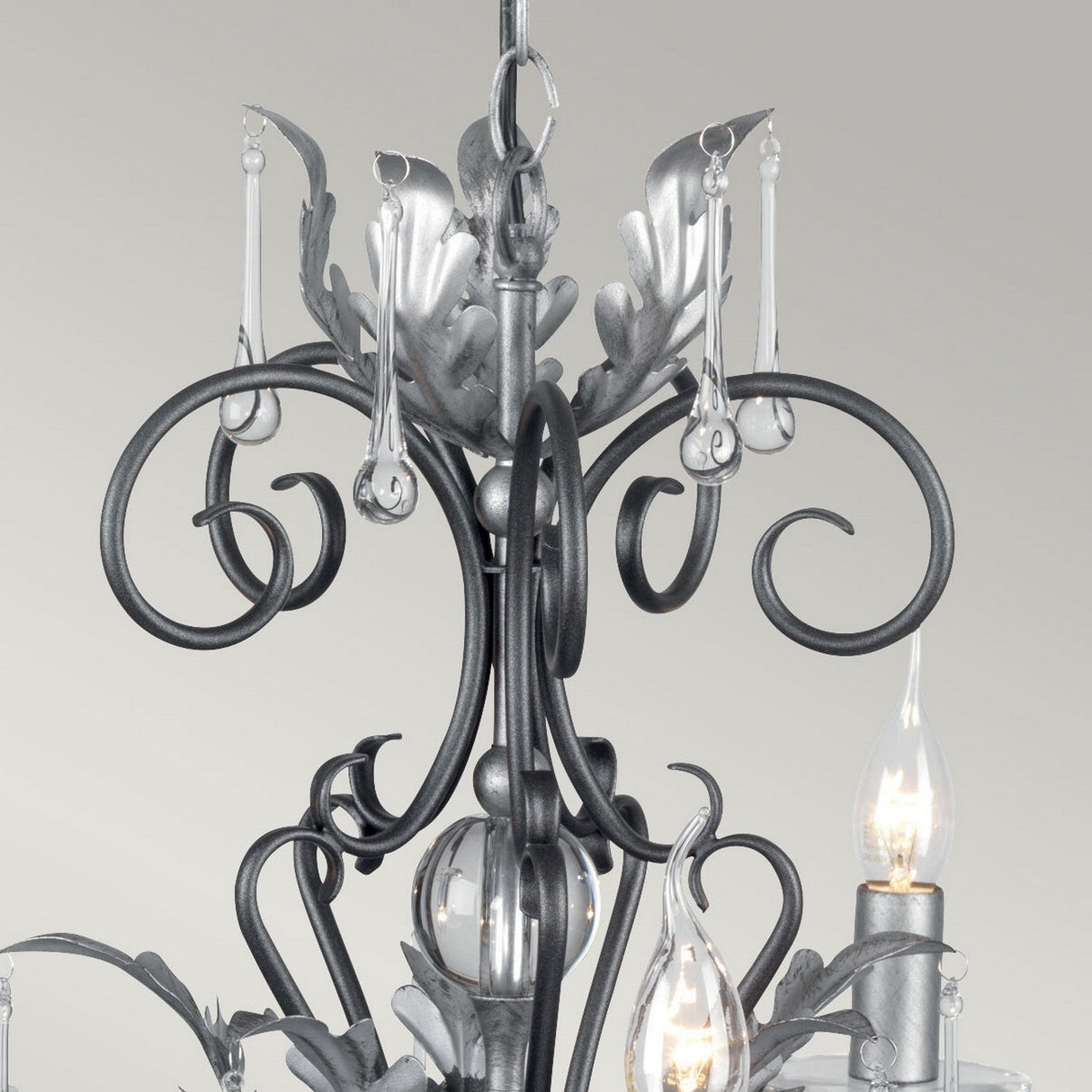Close-up of the Amarilli 5 Light Chandelier from Elstead, displaying its ornate design with intricate scrollwork and hanging glass ornaments. This chandelier boasts candle-shaped bulbs and a sophisticated silver finish complemented by a black patina, all set against a plain white background.