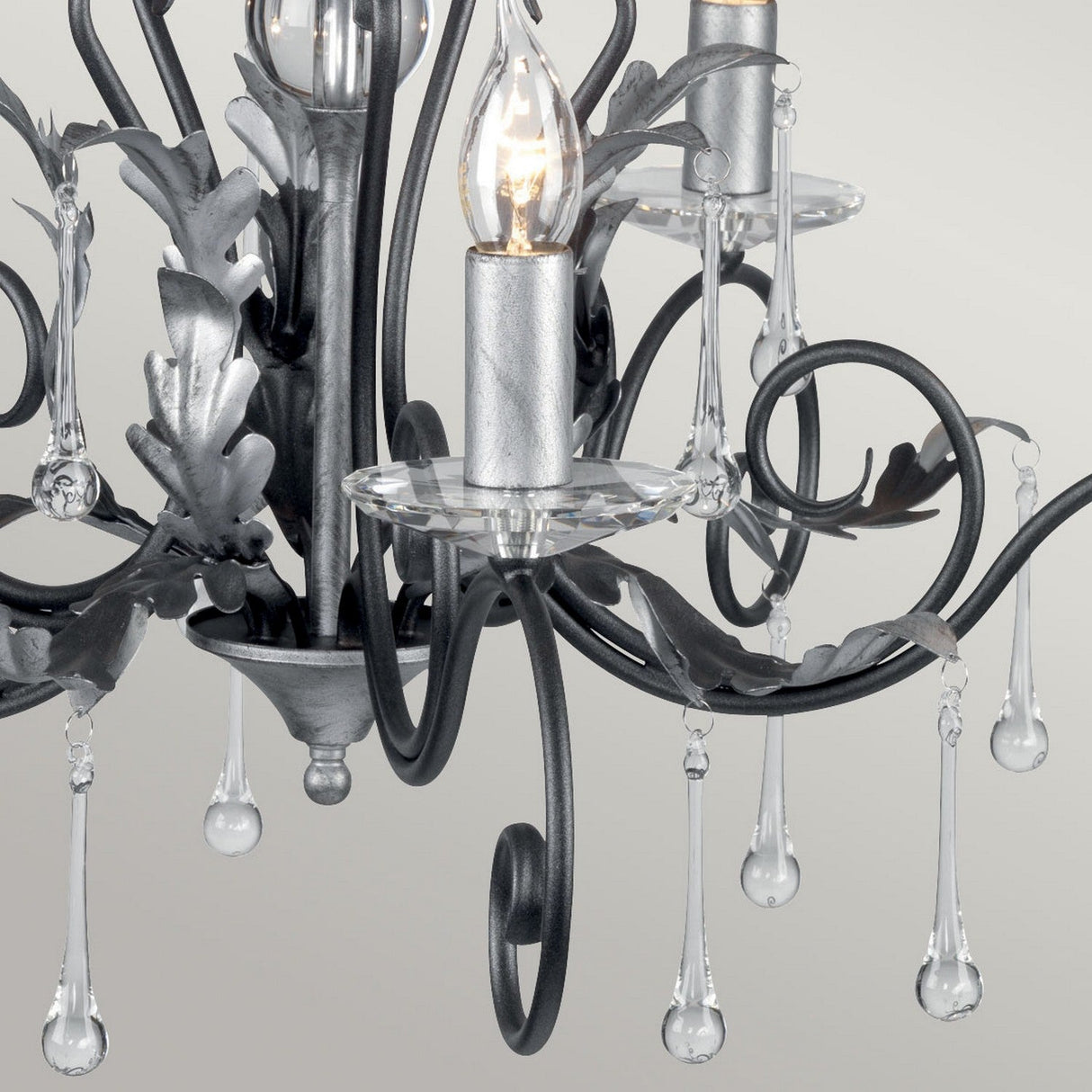 Close-up of the Amarilli 5 Light Chandelier in black with silver patina, showcasing intricate metal scrollwork and leaf designs. The chandelier features teardrop-shaped glass crystals and candle-like light bulbs, presented against a neutral background to highlight its elegance.