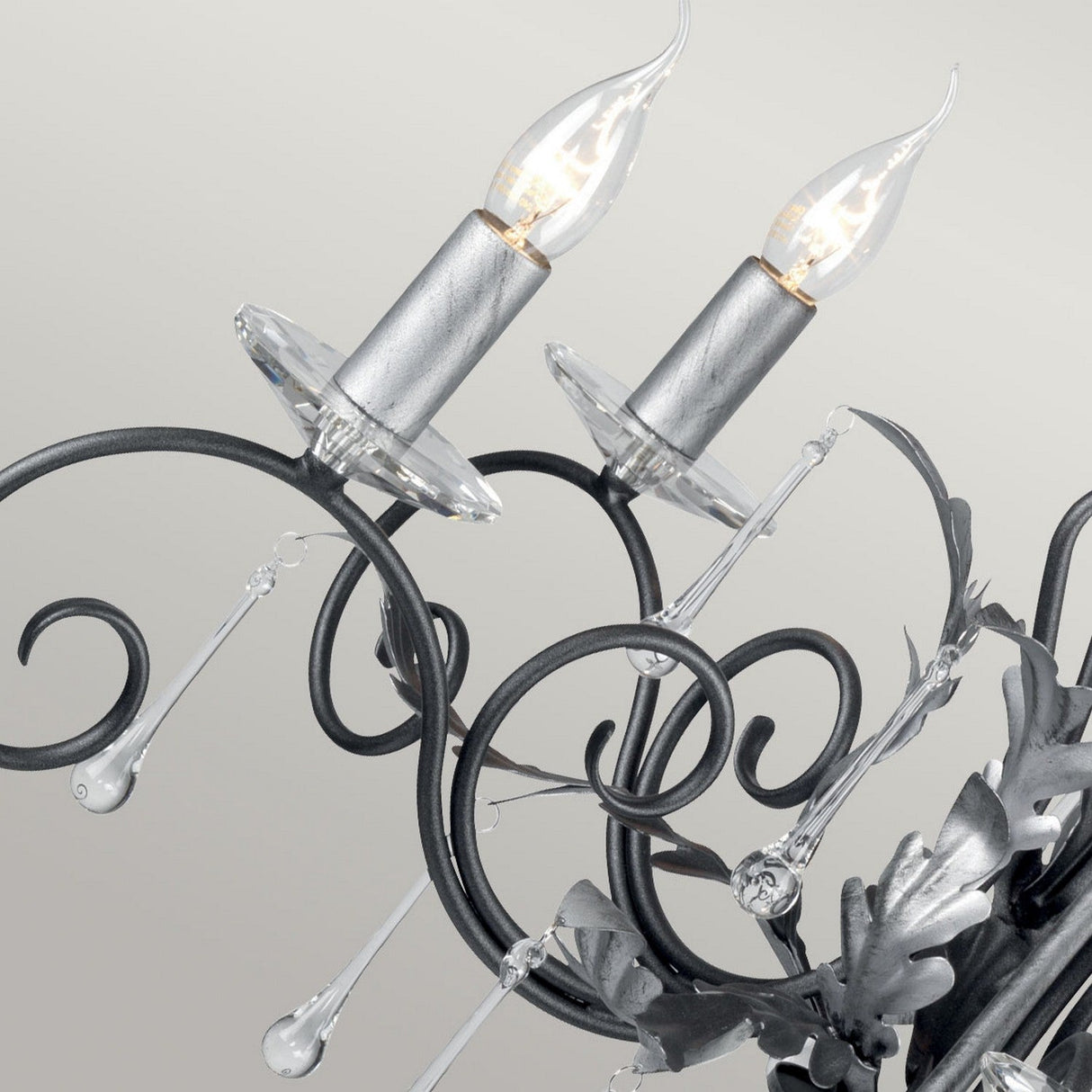 A close-up view of the Amarilli 5 Light Chandelier - Black With Silver Patina, showcasing its ornate vintage-style design. Two candle-shaped bulbs are lit, highlighting the fixture's intricate swirling metalwork in black with silver patina and adorned with clear crystal accents against a light gray background.