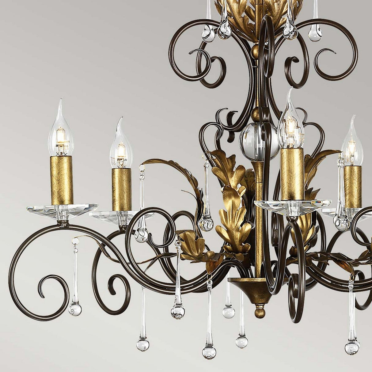 A close-up of the Amarilli 5 Light Chandelier - Bronze With Gold Patina showcases its ornate design, complete with bronze scrollwork, a gold patina finish, and five candle-shaped light bulbs adorned with crystal droplets, highlighting its elegant charm.