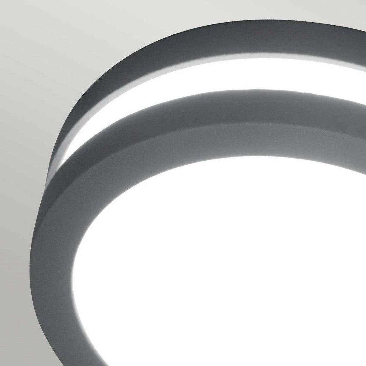 Close-up of the Ano-1 Outdoor Wall & Ceiling Light, featuring a modern round design with a sleek, cast aluminum frame in graphite finish. It emits a bright, even light against a light gray background, showcasing its minimalist aesthetic.