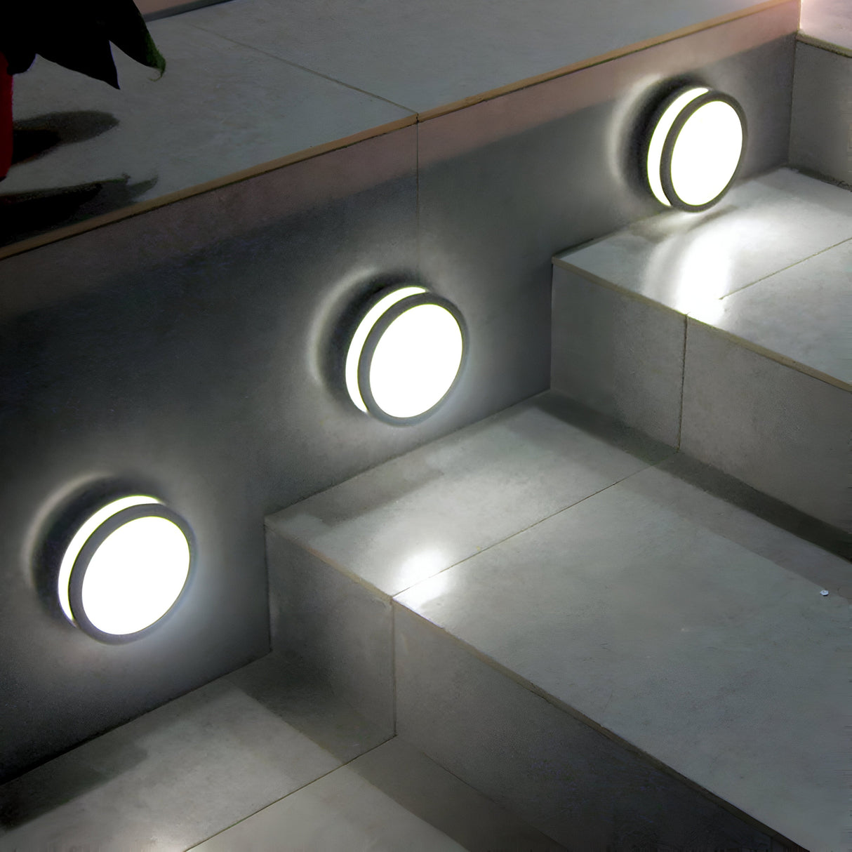 Three Ano-1 Outdoor Wall & Ceiling Lights in a graphite finish are embedded in a set of sleek, light-colored stone steps, casting a soft glow on the stairs and creating a contemporary and inviting ambiance.