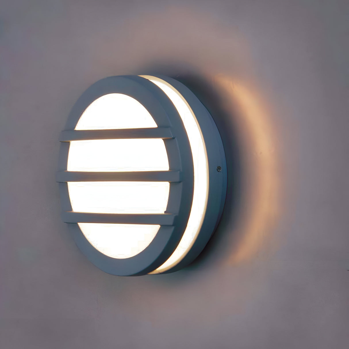 The Ano-2 Outdoor Wall & Ceiling Light in Graphite features a round, modern, and minimalist design that emits a warm glow. Its energy-efficient lighting showcases horizontal bars and a frosted cover, suitable for mounting on plain walls to create a subtle halo-like illumination ideal for outdoor settings.