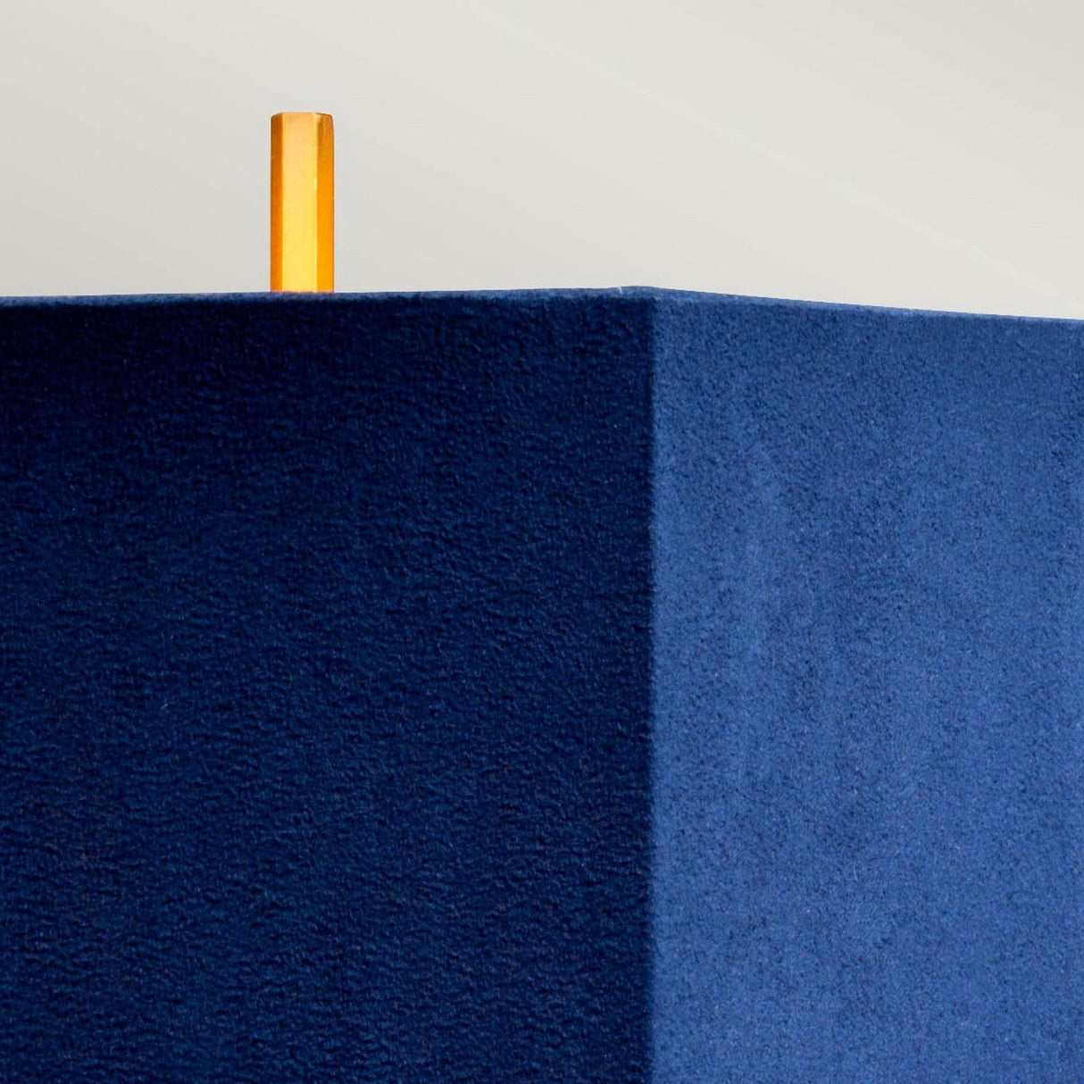 The image showcases the Apollo Table Lamp With Blue Shade - Brushed Brass, set against a light gray background. The design is minimalistic, featuring a textured blue cube-shaped shade with a small vertical cylinder in an orange hue that evokes the look of brushed brass protruding from the top, highlighting geometric shapes and contrasting colors.