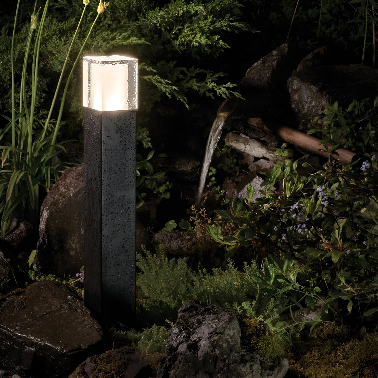 The Arendal 1 Light Large Bollard - Black provides a modern outdoor landscape lighting solution, beautifully illuminating a garden adorned with rocks, plants, and a small flowing water feature at night. This durable bollard enhances the lush greenery, creating a tranquil and natural setting perfect for coastal environments.