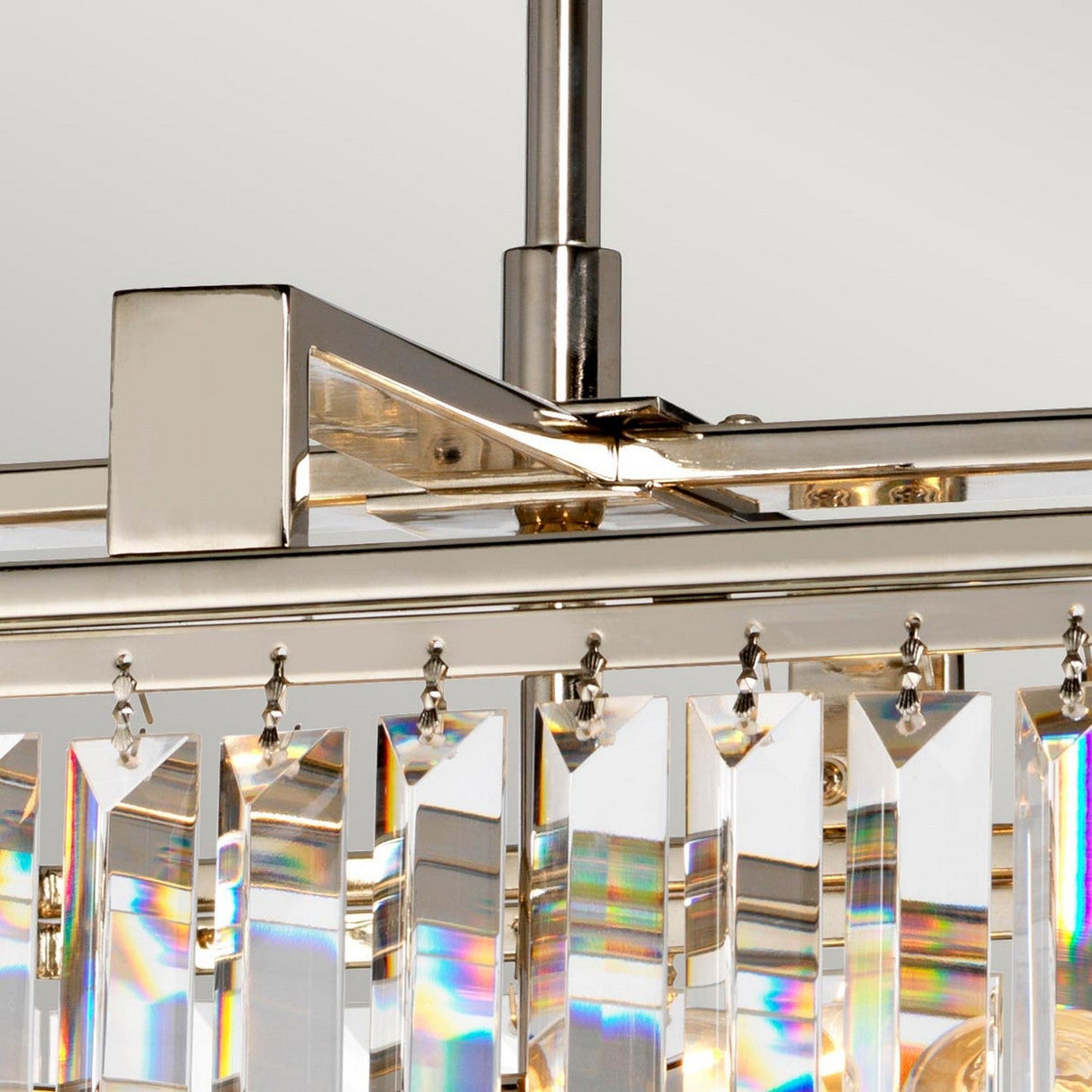 A detailed view of the Aries 4 Light Small Chandelier highlights its high-quality crystals, featuring rectangular, clear prisms that hang elegantly from a polished nickel metallic frame. These prisms reflect light beautifully, casting small rainbows as the fixture is suspended from a metal rod against a neutral background.