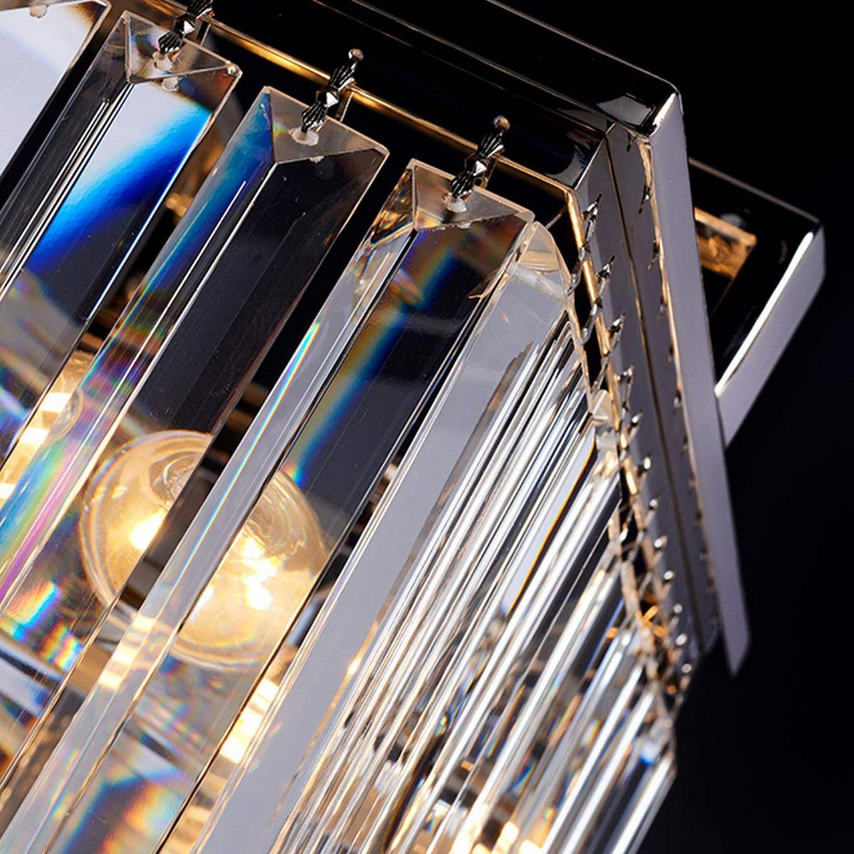 A close-up of the Aries 4 Light Small Chandelier in polished nickel showcases its high-quality crystal prisms. The polished nickel accents amplify the rainbow effect as they reflect light from the bulbs within, set against a dark background.