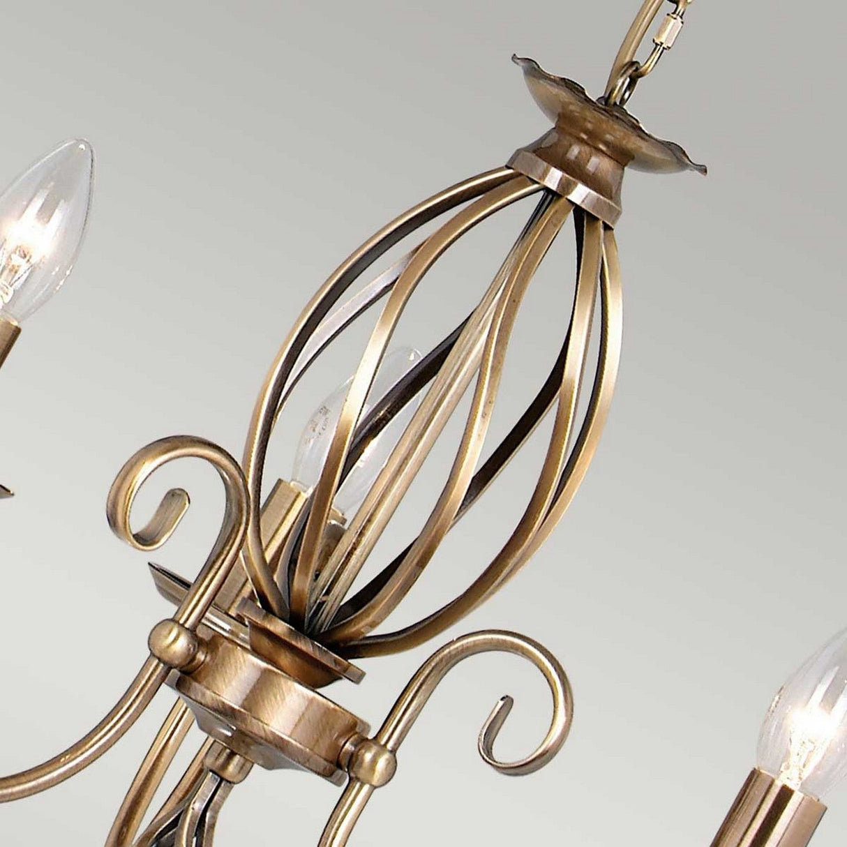 A close-up of the Artisan 3 Light Chandelier - Aged Brass showcases its decorative curling arms and candle-style bulbs. The elegant design is enhanced by an open metal cage centerpiece, and it offers a flush convert option for versatile installation.