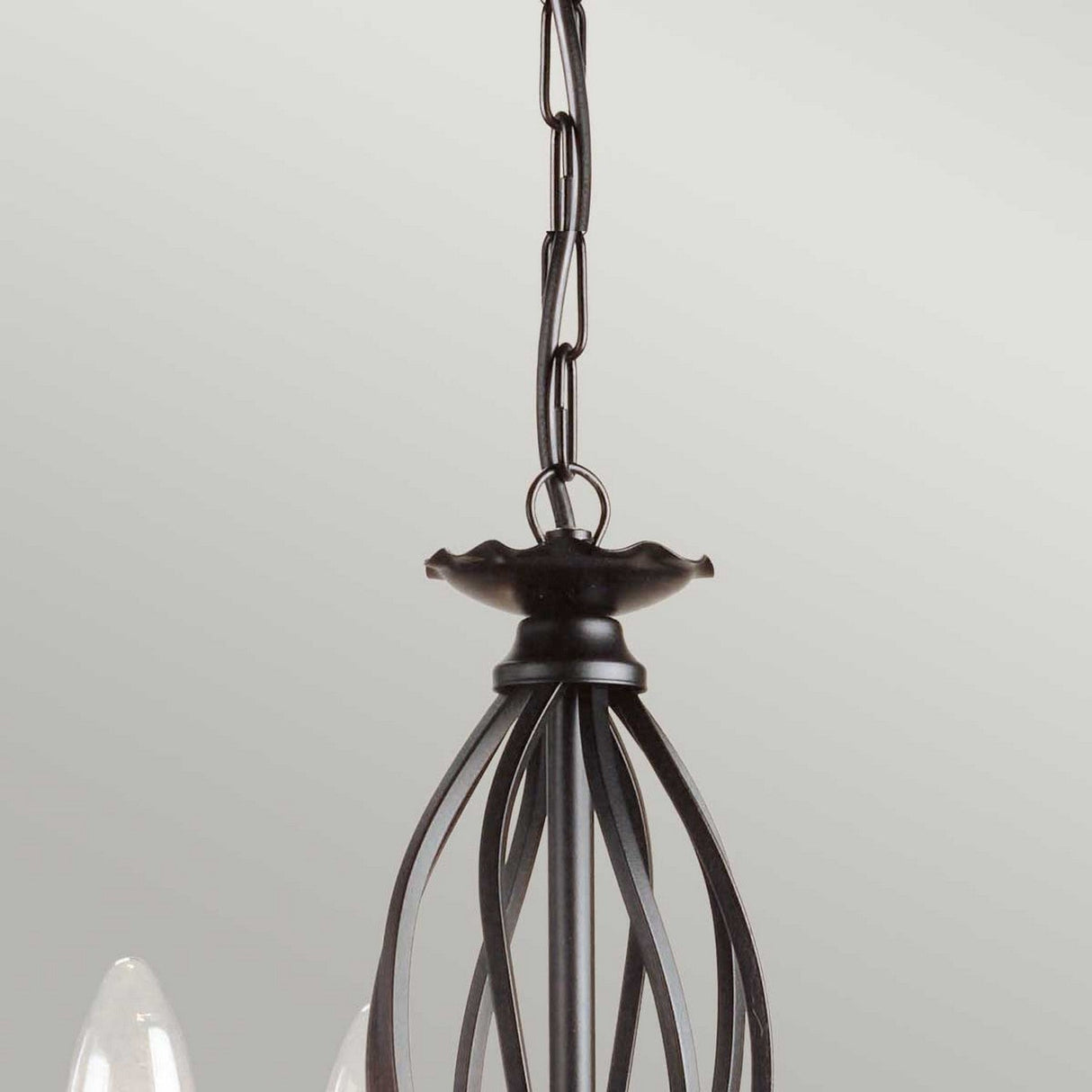 The Artisan 3 Light Chandelier - Black exudes graceful ambient lighting with sleek, curved metal rods and a refined black finish. Hanging from a chain, the chandelier highlights a visible bulb set against a plain, light-colored wall, introducing an artistic element to any space.