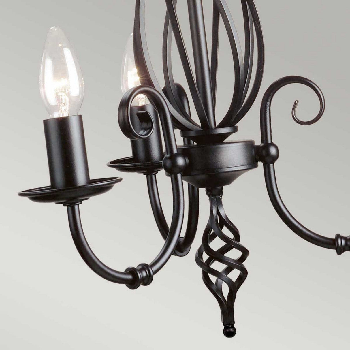 A close-up of the Artisan 3 Light Chandelier - Black reveals intricate scrollwork in black metal and features two unshaded light bulbs. The craftsmanship emphasizes the elegant curves and artistic details, providing ambient lighting against a plain background.