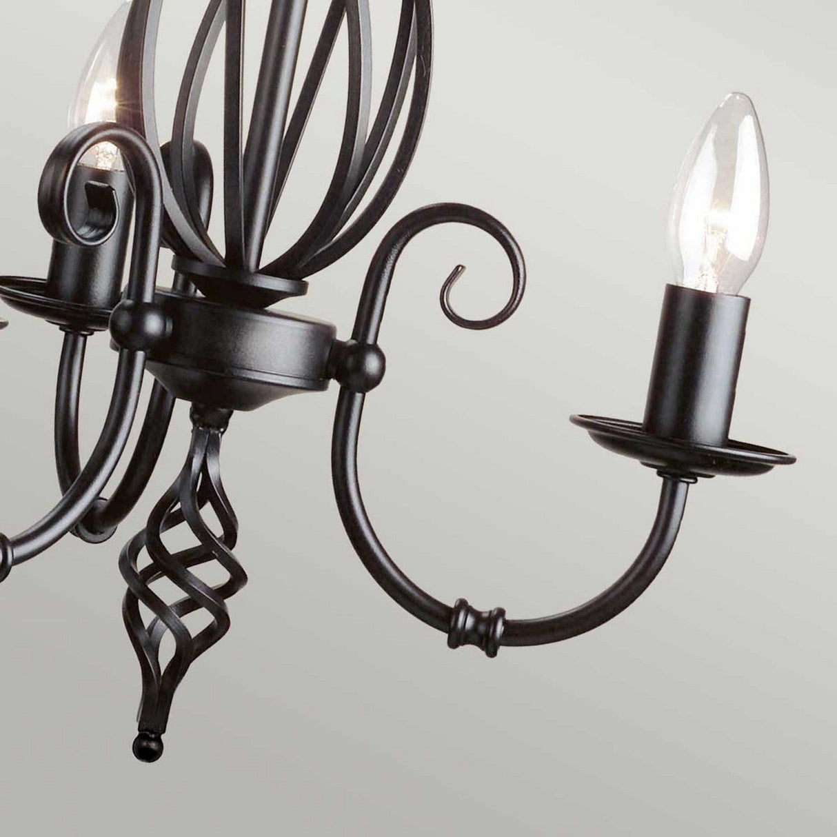 Close-up of the Artisan 3 Light Chandelier - Black, an intricate metal fixture showcasing two glowing candle-shaped bulbs. It boasts elegant, swirling arms and a twisted central column, ideal for creating ambient lighting against a simple backdrop.