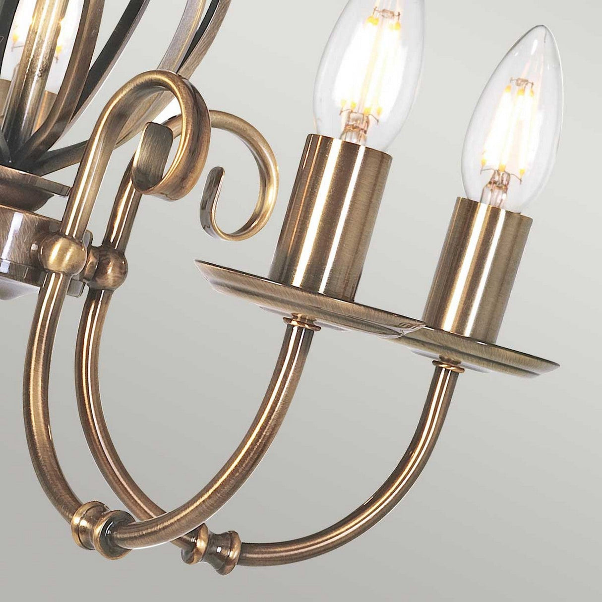Close-up of the Artisan 5 Light Chandelier in aged brass, showcasing two lit candle-style LED bulbs. This decorative focal point features elegantly curved metal arms and a classic design, set against a neutral background.