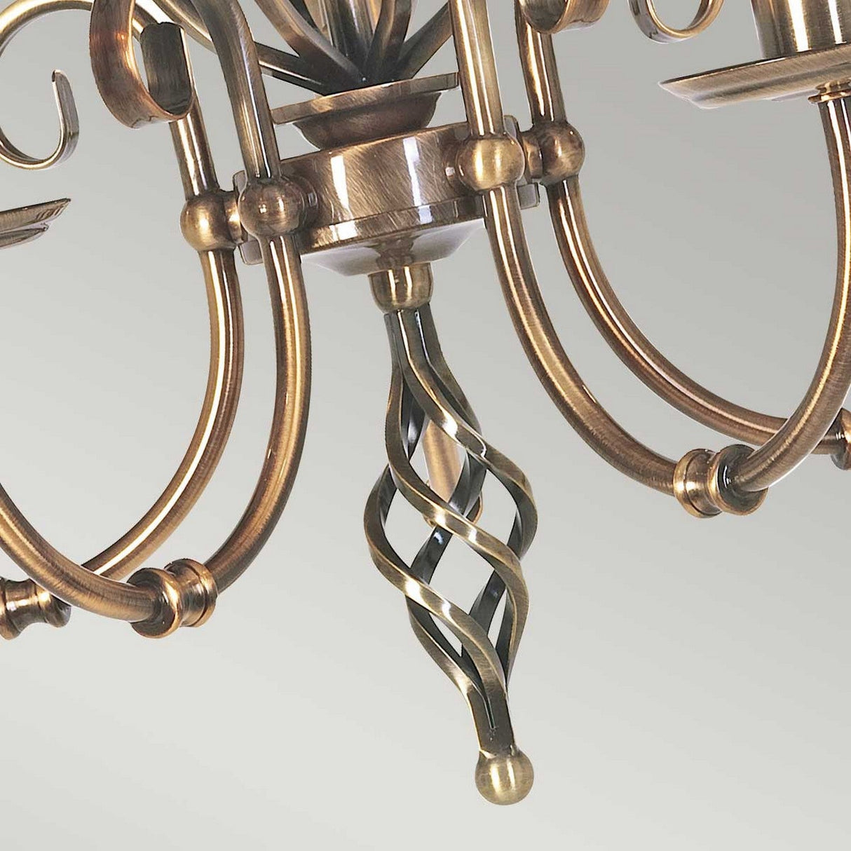A close-up of the Artisan 5 Light Chandelier in aged brass showcases its twisted central rod and multiple elegantly curved arms. The chandelier's shiny finish beautifully reflects light, making it a stunning decorative focal point.