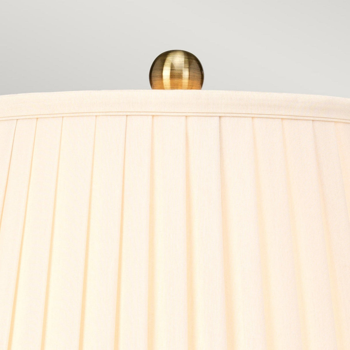 Close-up of the top of a beige pleated lampshade with an aged brass finial, reminiscent of the Arum Table Lamp - Aged Brass. The background is a plain, light gray.