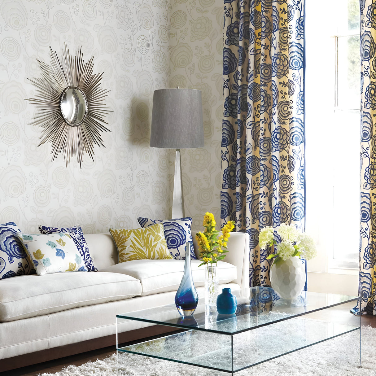 A modern living room with a white sofa and colorful floral cushions. A glass coffee table is adorned with blue vases and yellow flowers. The wall features a decorative mirror beside the Ascent Floor Lamp With Dark Grey Shade in a polished nickel finish. Blue and yellow patterned curtains frame the window.