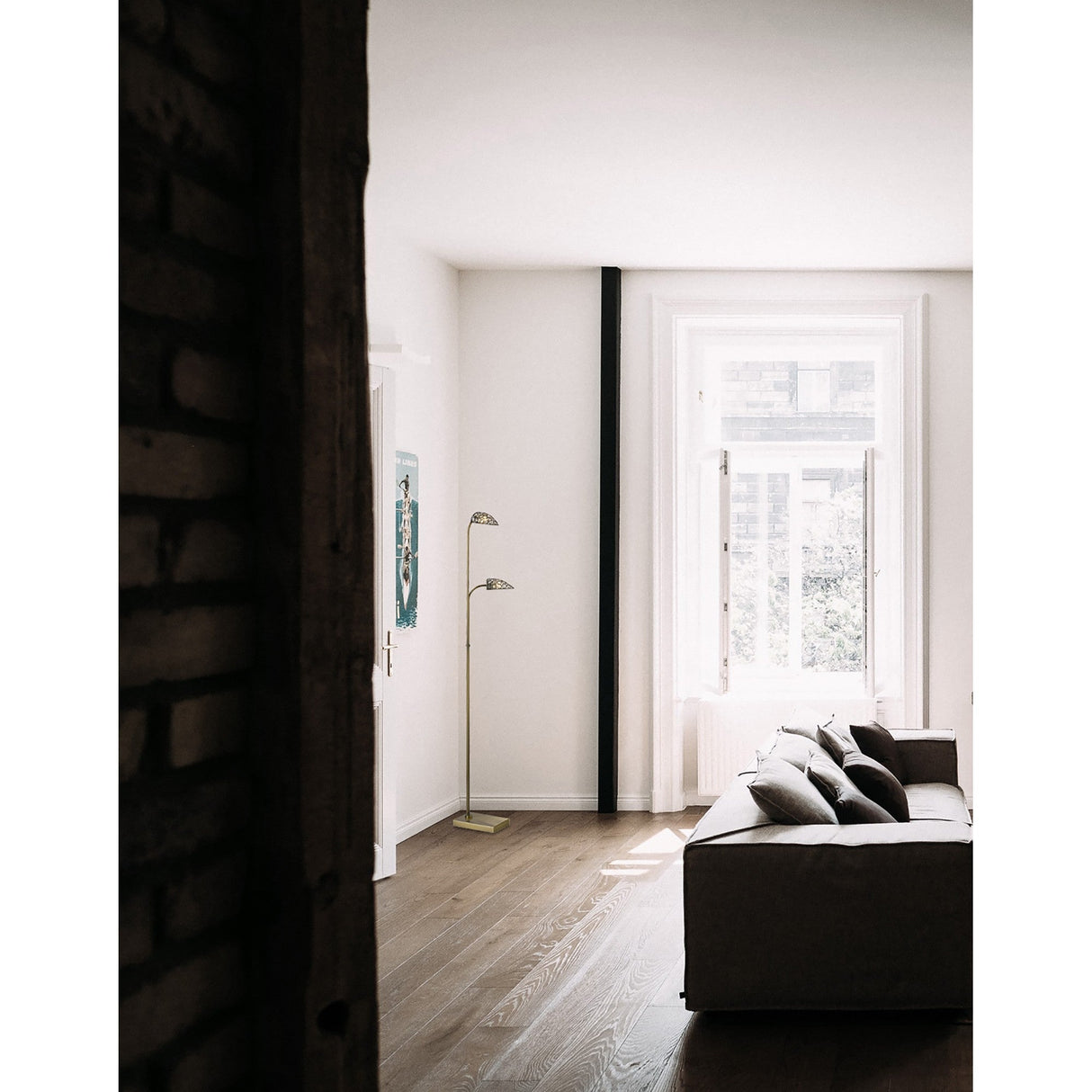 A bright, minimalist living room features a gray sofa, wooden floors, a large window, and ambient lighting from two Ashton 2 Light Crystal Floor Lamps in Antique Brass. Part of a brick wall is visible in the left foreground.