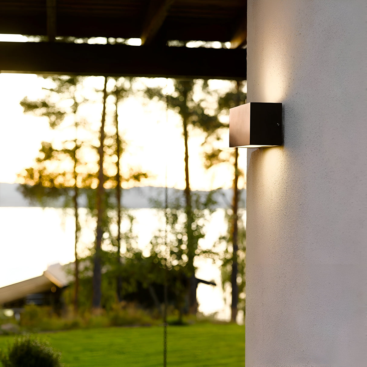 The Asker Outdoor 1 Light Up/Down in graphite, featuring an IP65 rating, is mounted on the building's exterior, casting a warm glow as dusk falls. In the background, a serene landscape with trees and water can be seen beneath the setting sun on the horizon.