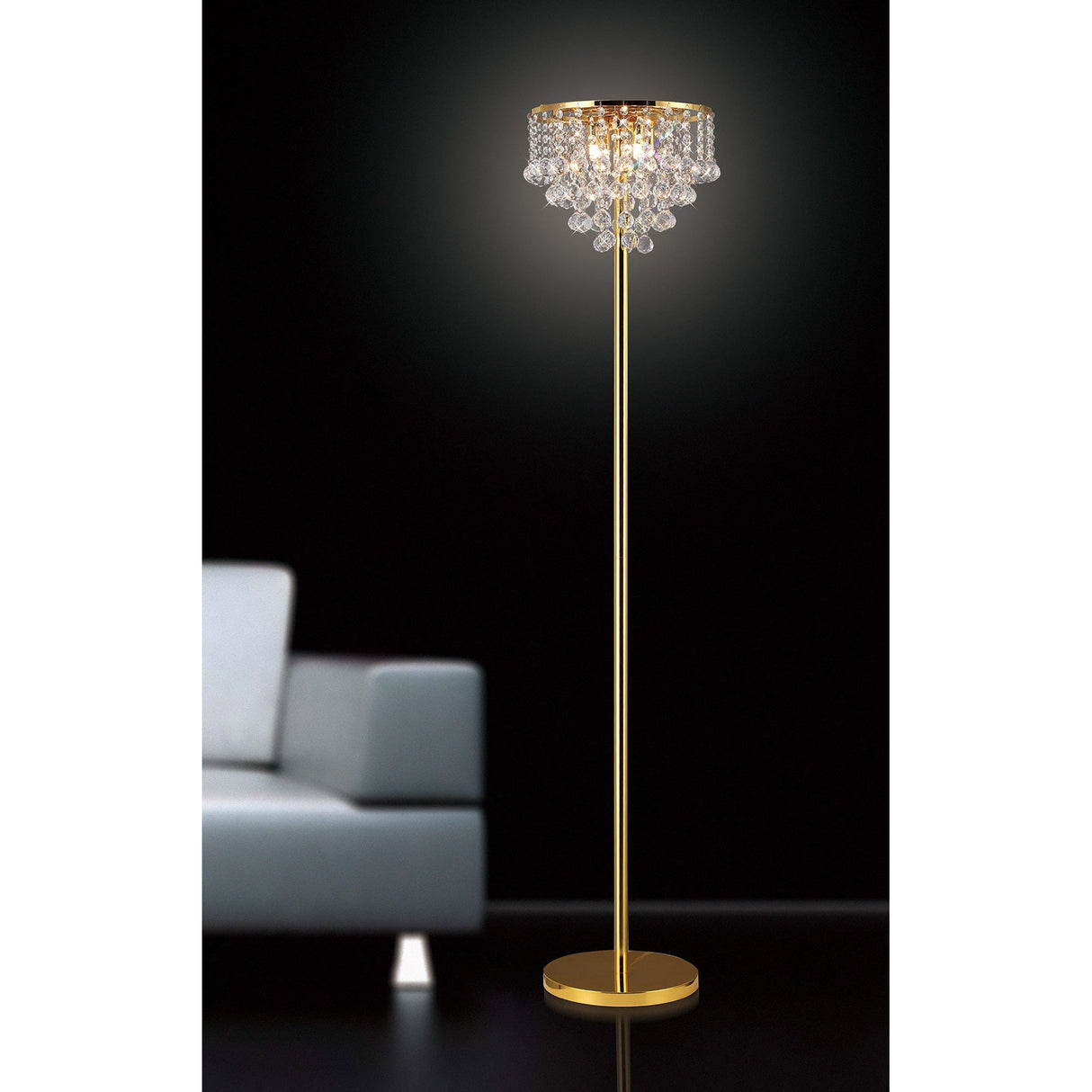 The Atla 4 Light Crystal Floor Lamp in French Gold stands illuminated on a black background beside a light gray modern sofa, its chandelier-style shade casting a warm glow.