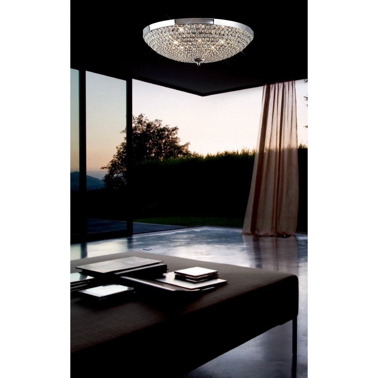 Diyas Ava 6 light crystal flush ceiling light in polished chrome, illuminated above a dark modern living space with floor-to-ceiling windows at dusk.