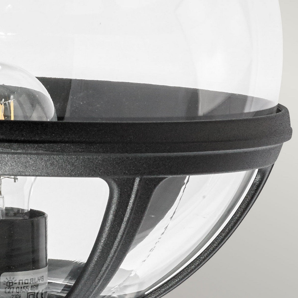 A close-up view showcases the Bologna Outdoor 1 Light Up Wall Light in black, featuring a clear glass bulb encased in a spherical black metal cage. The elegant design reveals the bulb's filament and base against a neutral, light gray background.