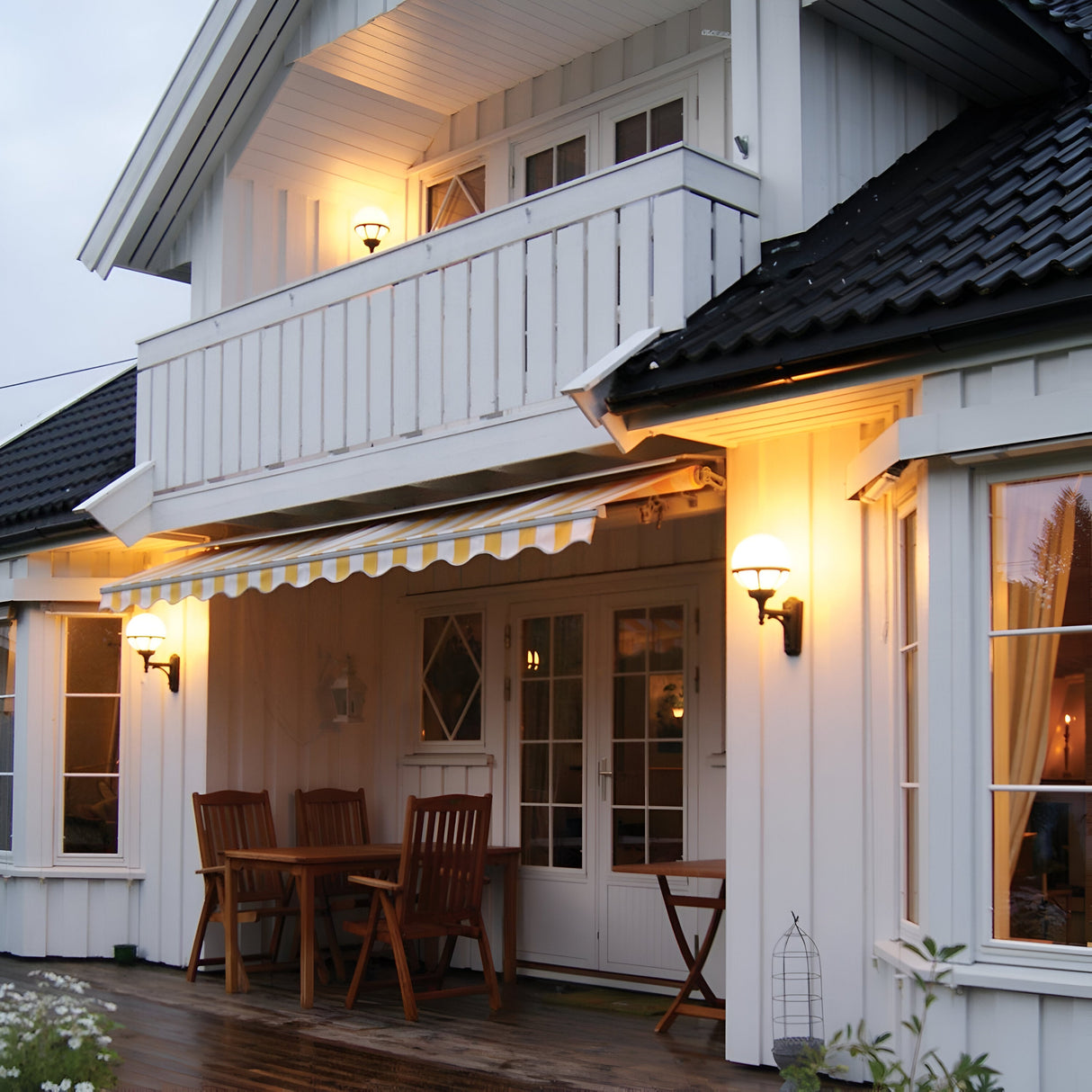 A cozy white house with a balcony features the Bologna Outdoor 1 Light Up Wall Light in black, casting a warm glow. A striped awning extends over a wooden patio furnished with a table and chairs. Inside, dimmable lamps reflect off the windows, creating a welcoming evening atmosphere.