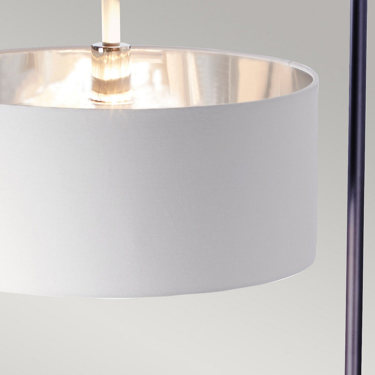The image features a close-up of the Balance Floor Lamp - Black/Polished Nickel, highlighting its white fabric drum shade with a sleek polished nickel interior. It is mounted on a slender vertical metal rod. When lit, the lamp emits an energy-efficient glow that enhances the polished surface. The background is plain and light gray, complementing modern decor seamlessly.