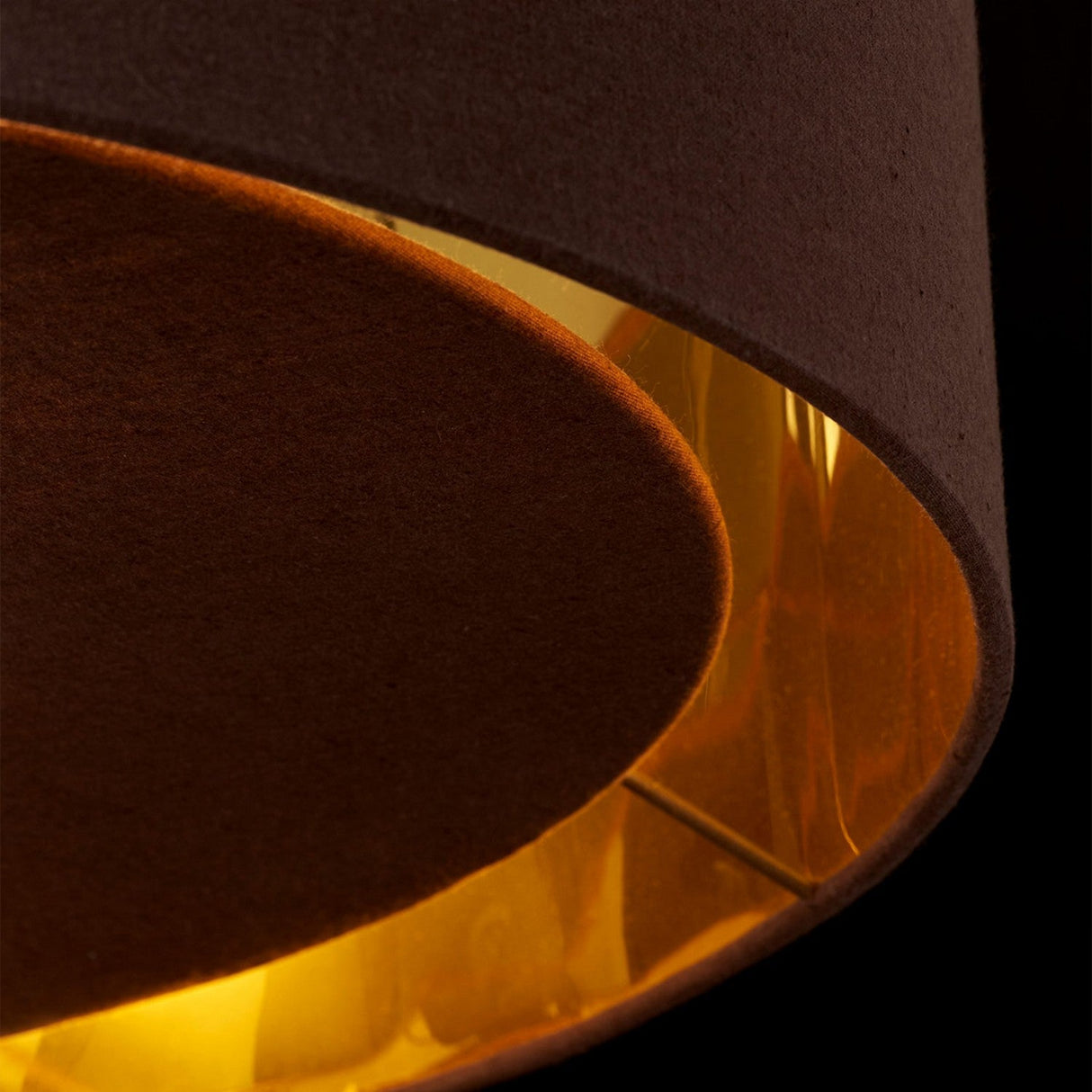 Close-up of the Balance Pendant Light - Brown/Polished Brass, featuring a sleek brown exterior and a warmly glowing interior. Its energy-efficient lighting creates a cozy ambiance, emphasizing the textured fabric. The dark background enhances the illumination of the lamp.
