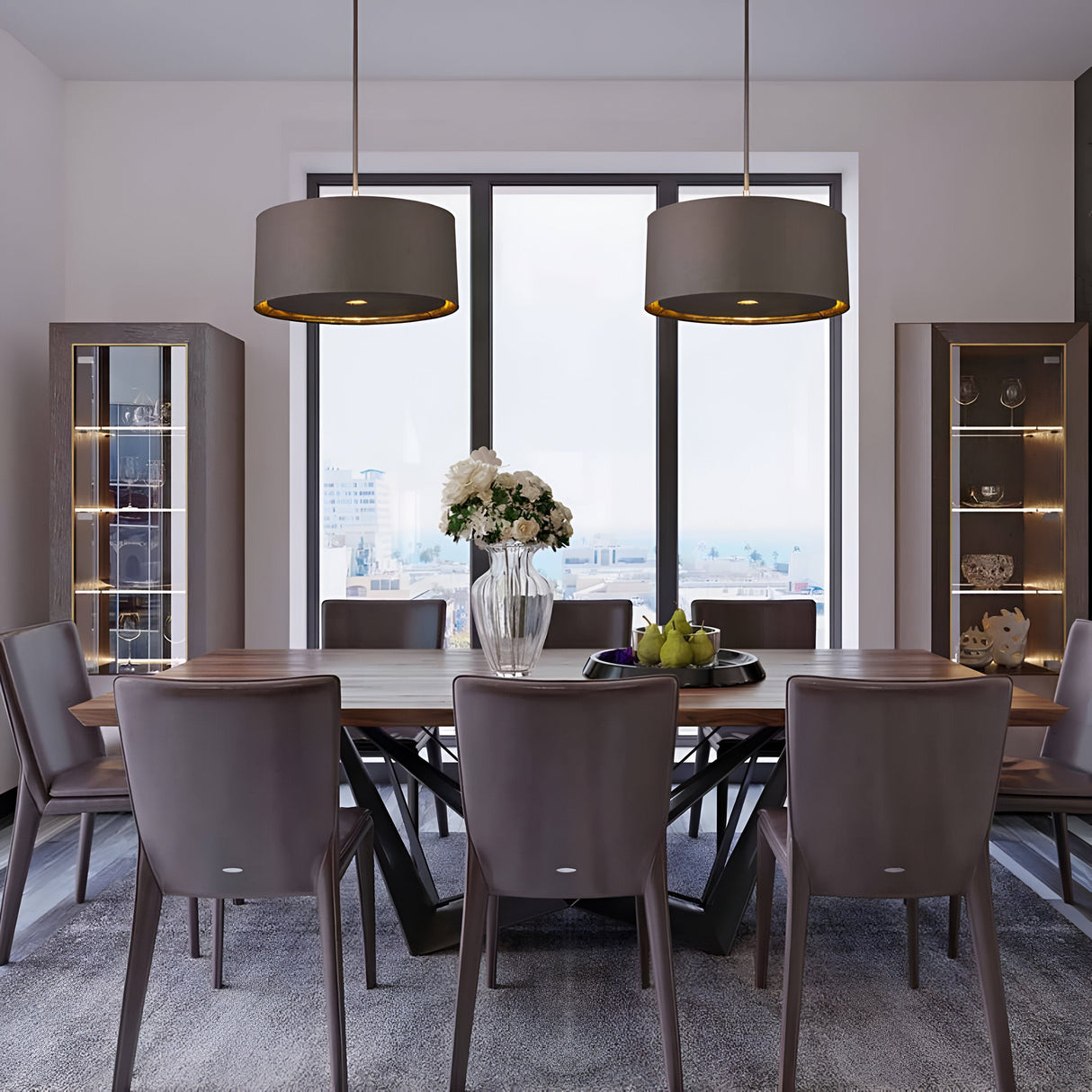 A modern dining room features a wooden table surrounded by eight gray chairs. Above the table, two Balance Pendant Lights in brown and polished brass illuminate a vase of white flowers and a bowl of green apples. Tall glass cabinets flank a large window with a city view, enhancing the setting's elegant ambiance.