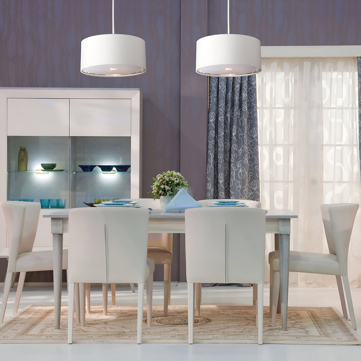 A modern dining room features a light wood table, six white chairs, and two Balance Pendant Lights in White/Polished Nickel providing energy-efficient illumination. A cabinet with glass dishes and a vase of flowers complement the curtains with blue and white patterns, enhancing the elegant atmosphere.
