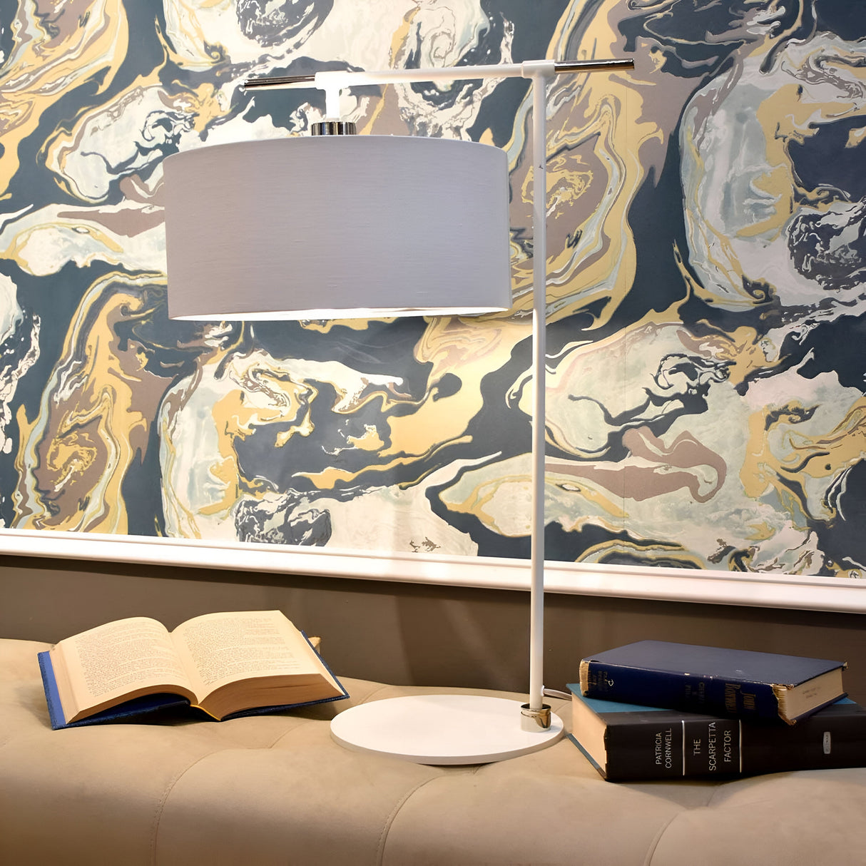 The Balance 1 Light Table Lamp in white and polished nickel sits elegantly on a table, featuring its sleek design with a cylindrical shade. An open book lies nearby, accompanied by two closed books stacked beside it. The background showcases a colorful, swirling marble-patterned wallpaper that enhances the ambiance with its modern flair.
