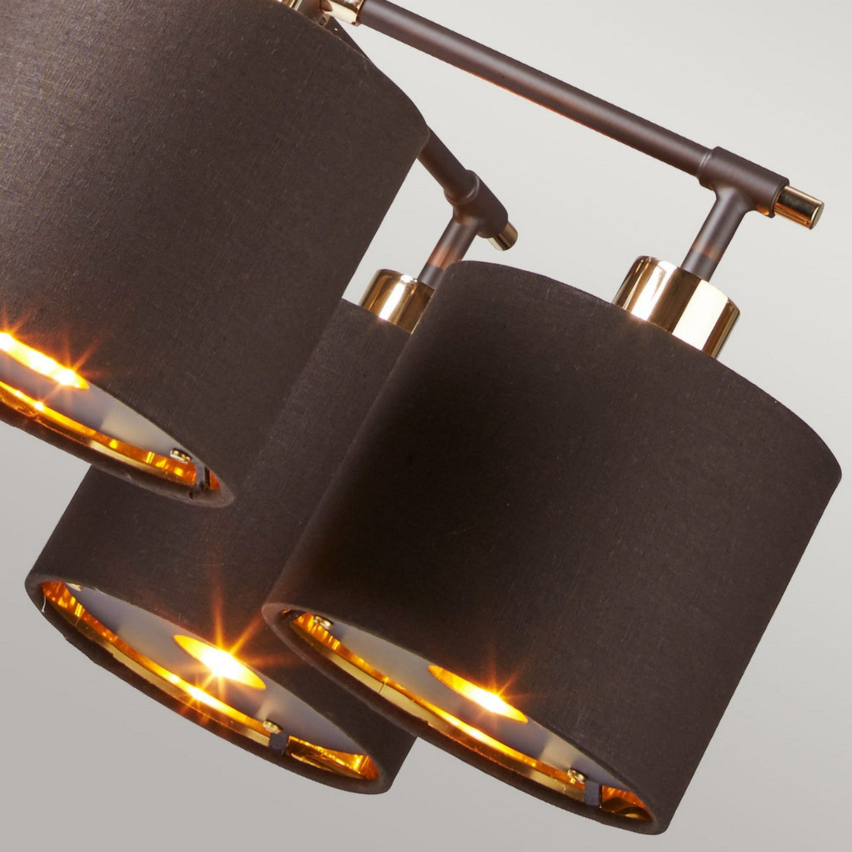 A close-up of the Balance 5 Light Chandelier showcases its modern design with three cylindrical shades featuring a dark fabric exterior. The lights inside cast a warm glow, perfect for creating a cozy ambiance. Its sleek design is highlighted by a polished brass finish, seamlessly combining style with energy-efficient lighting.