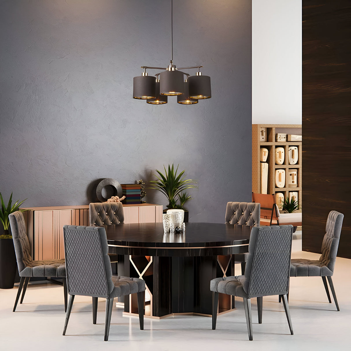 A modern dining room showcases a round black table paired with six quilted gray chairs, elegantly illuminated by the Balance 5 Light Chandelier in brown/polished brass mounted above. The space is accented with decorative plants and artifacts on a sideboard, complemented by bookshelves in the background.