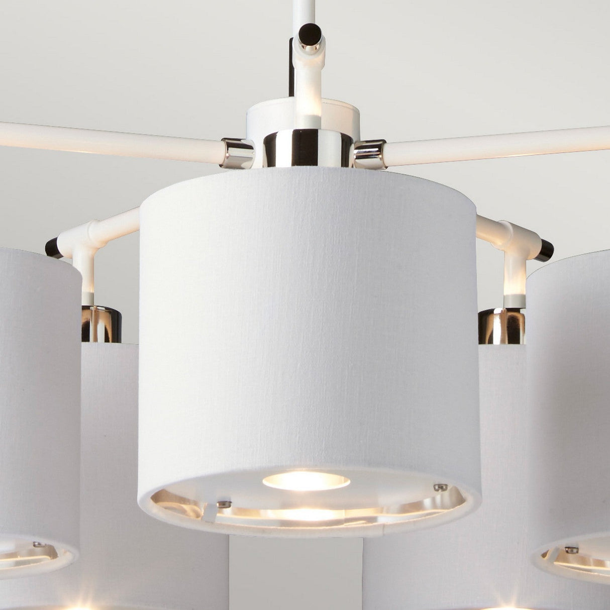 Close-up of the Balance 5 Light Chandelier in white with polished nickel accents. The modern cylindrical fixture showcases a smooth fabric texture and features visible light bulbs at the bottom, set against a plain white ceiling.