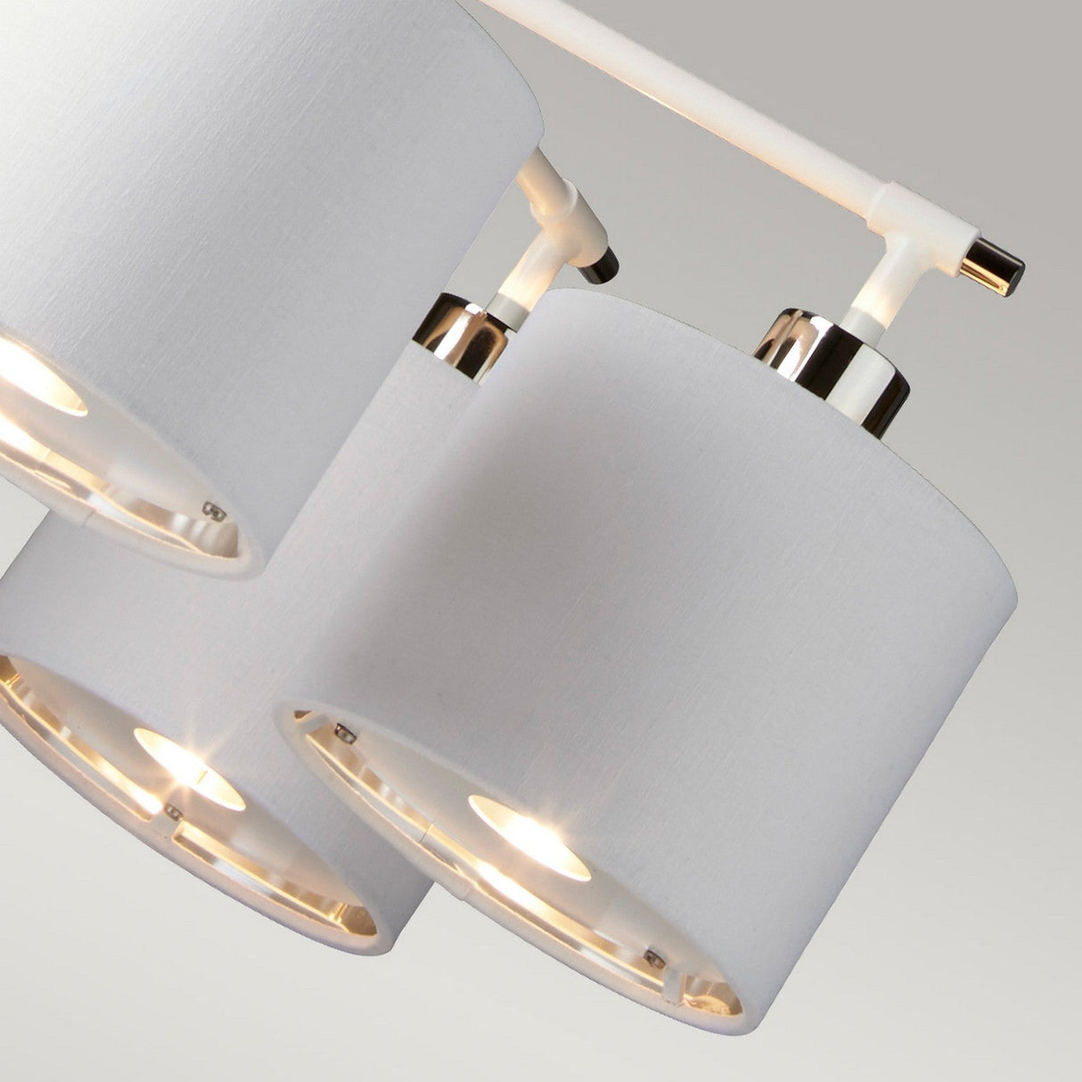 A close-up of the Balance 5 Light Chandelier in White/Polished Nickel features three modern pendant lights with white cylindrical fabric shades and polished metallic accents, each emitting a warm, energy-efficient glow. The chandelier is suspended from a sleek white rod against a plain background, highlighting its contemporary design.