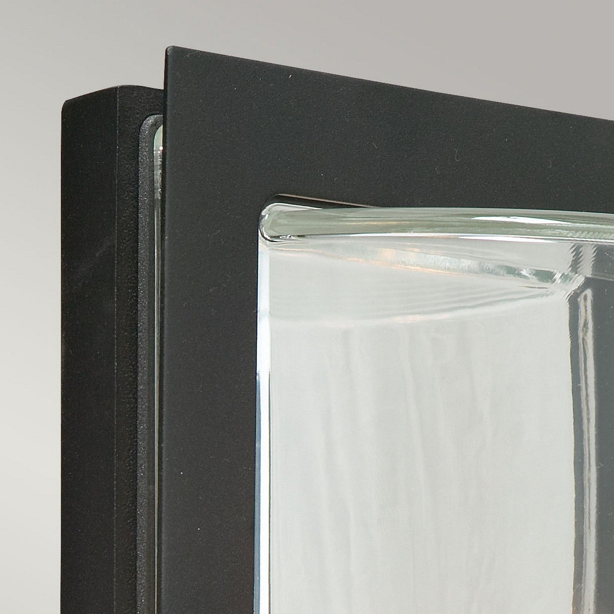 A detailed view of the corner of a glass block with a clear, wavy texture framed in a sleek black finish. The Basel Outdoor 1 Light Wall Lantern exudes modern elegance against a plain, light gray background.