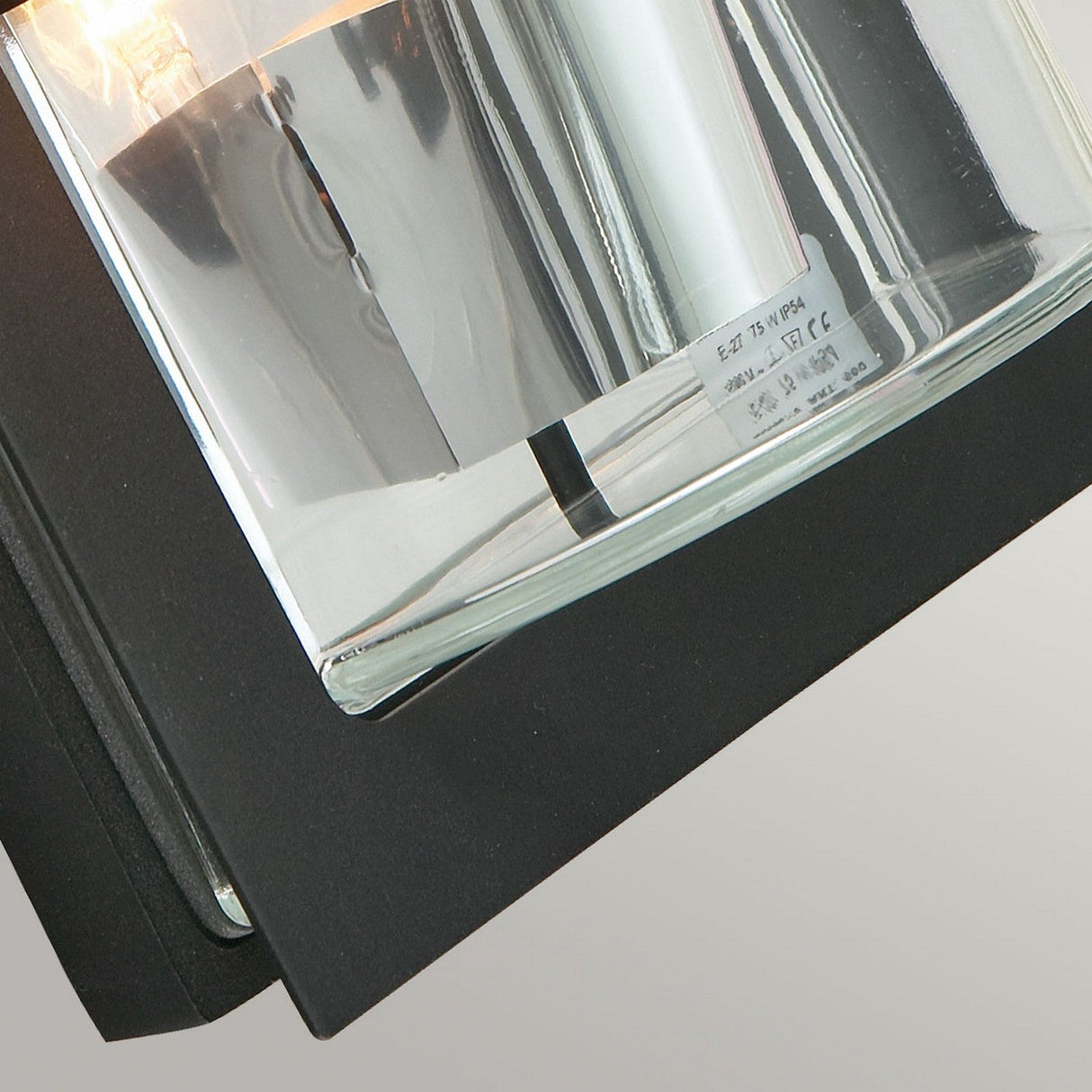 Close-up of the Basel Outdoor 1 Light Wall Lantern, featuring a sleek black finish and a rectangular clear glass cover. The light is illuminated, casting a warm glow through the transparent glass.